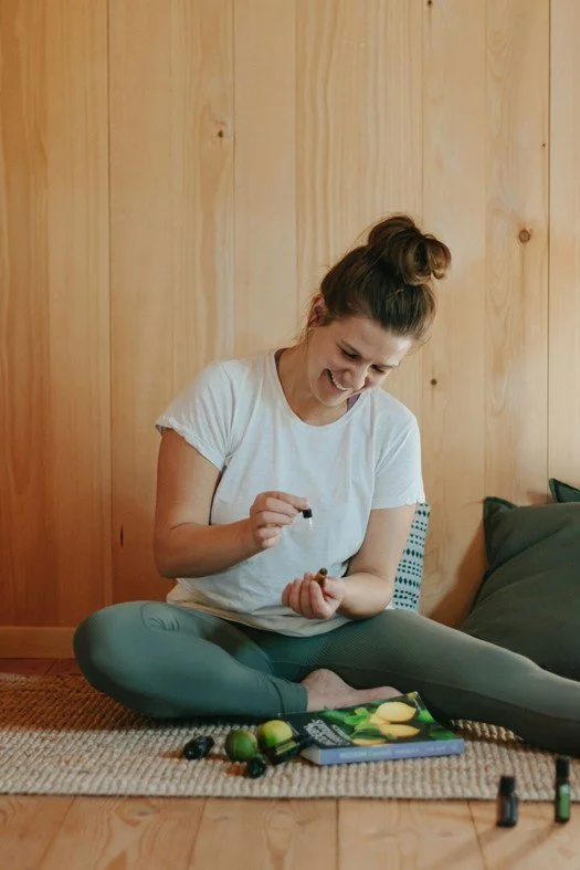 Femme assise par terre appliquant de l'huile essentielle, portant un t-shirt blanc et un leggings vert, avec des flacons d'huiles essentielles sur le tapis devant elle, un mur en bois à l'arrière-plan.