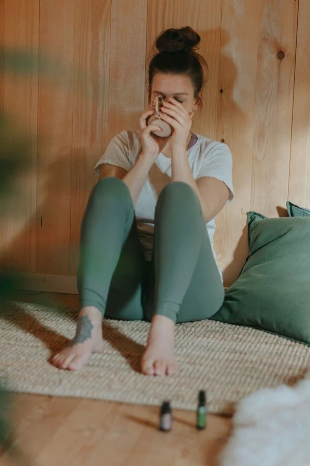 Femme assise sur un tapis, tenant une tasse, portant un t-shirt blanc et un pantalon vert, avec des flacons d'huiles essentielles au sol.