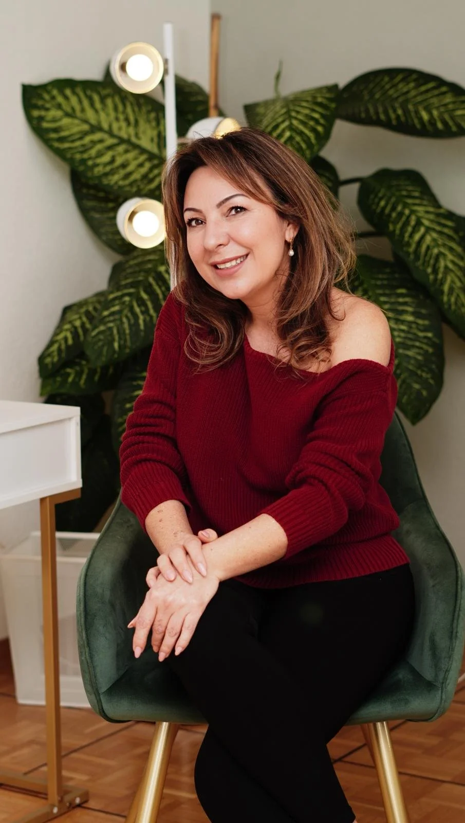 Femme souriante assise sur une chaise, portant un pull rouge, avec des plantes en arrière-plan.