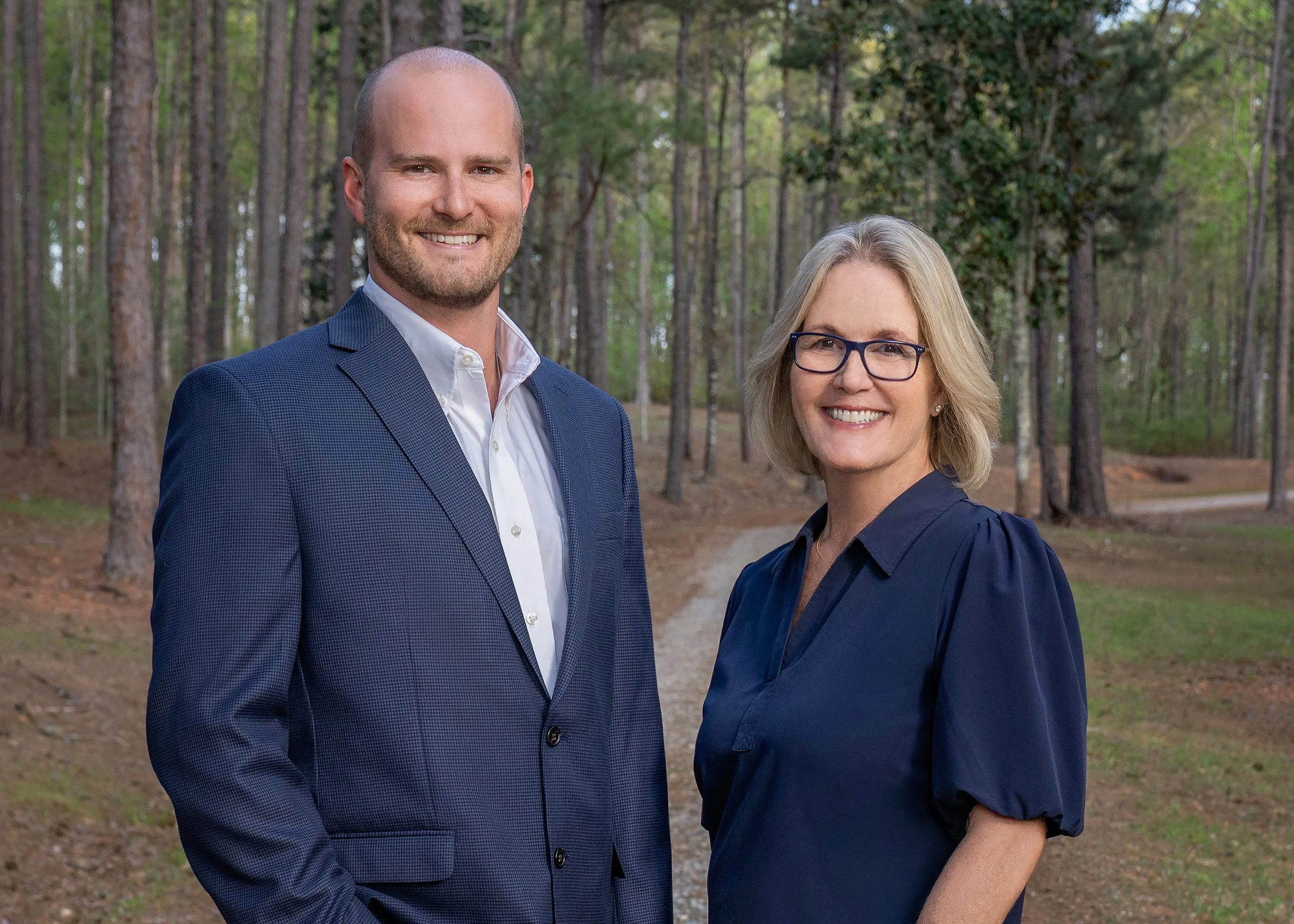 A man and a woman standing outdoors in a wooded area, smiling at the camera.