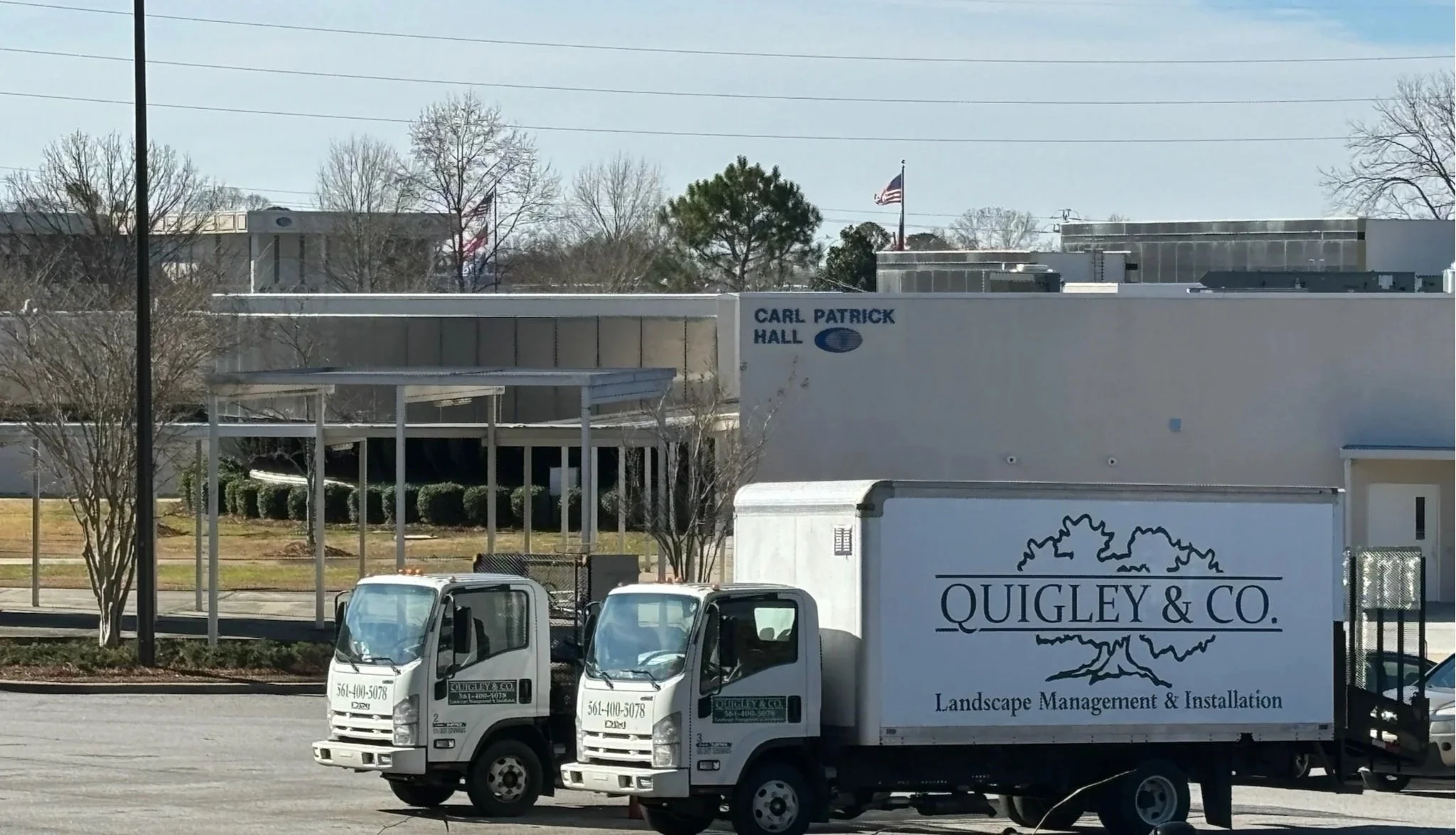 Two landscaping trucks from Quigley & Co. parked on a street in front of a building with a sign that reads Carl Patrick Hall; the trucks and building are surrounded by trees and American flags.