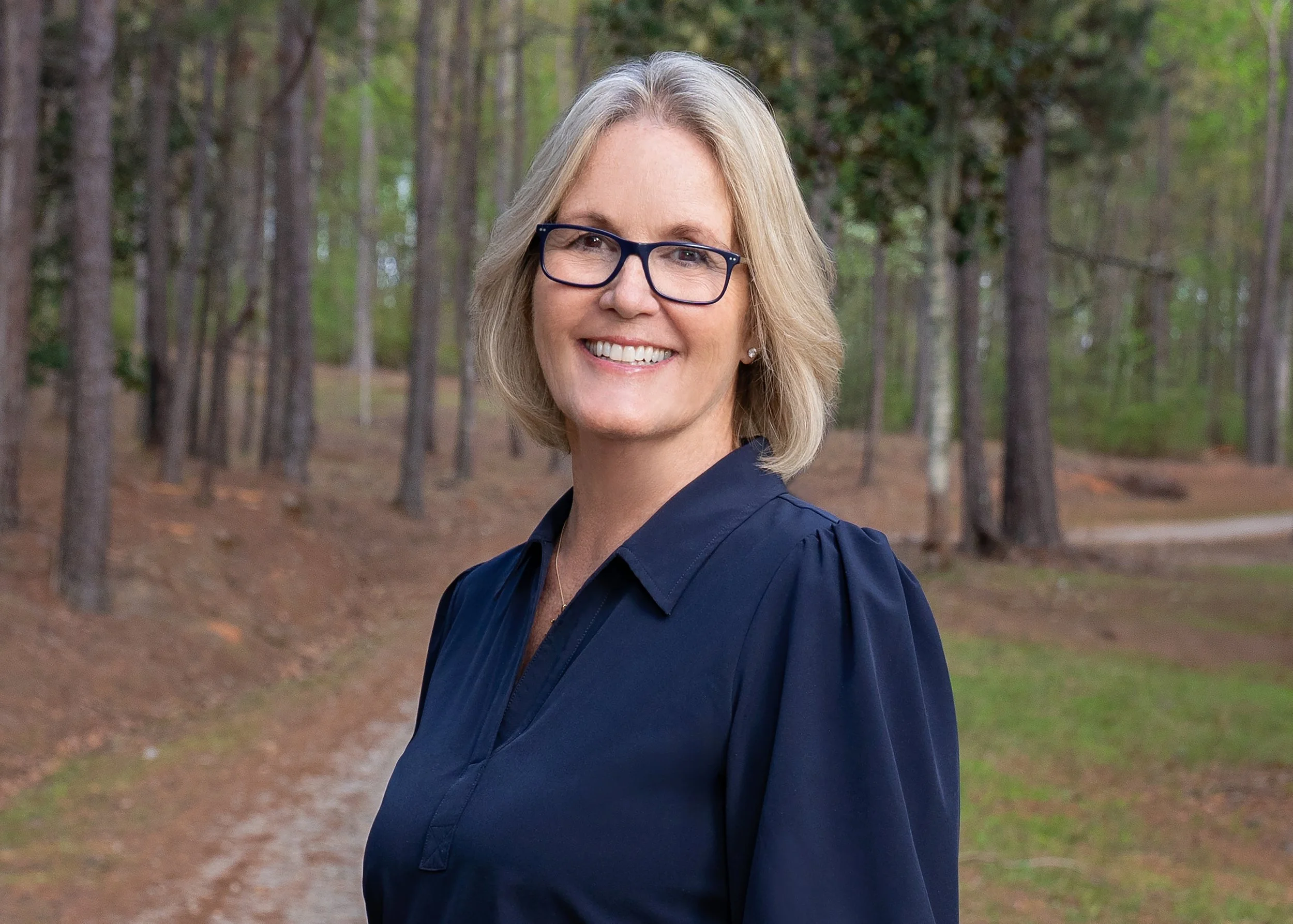 A smiling woman with blonde hair, glasses, and a navy blouse standing outdoors in a wooded area with trees and a dirt path in the background.