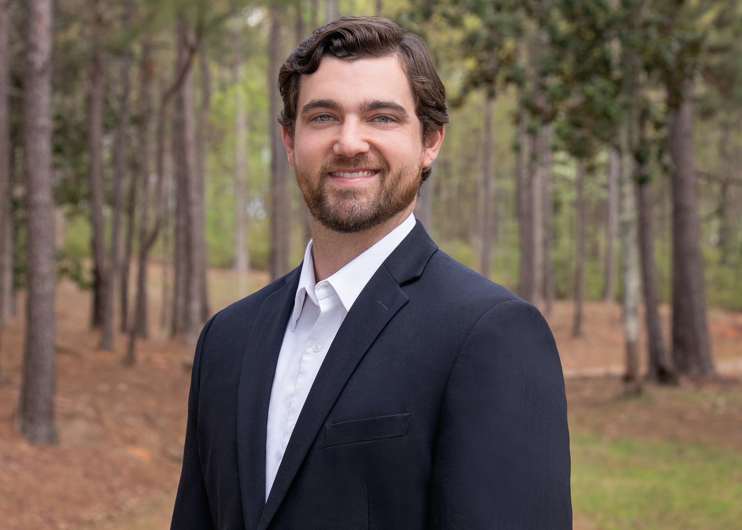 A man in a navy suit and white shirt standing outdoors in a wooded area, smiling at the camera.