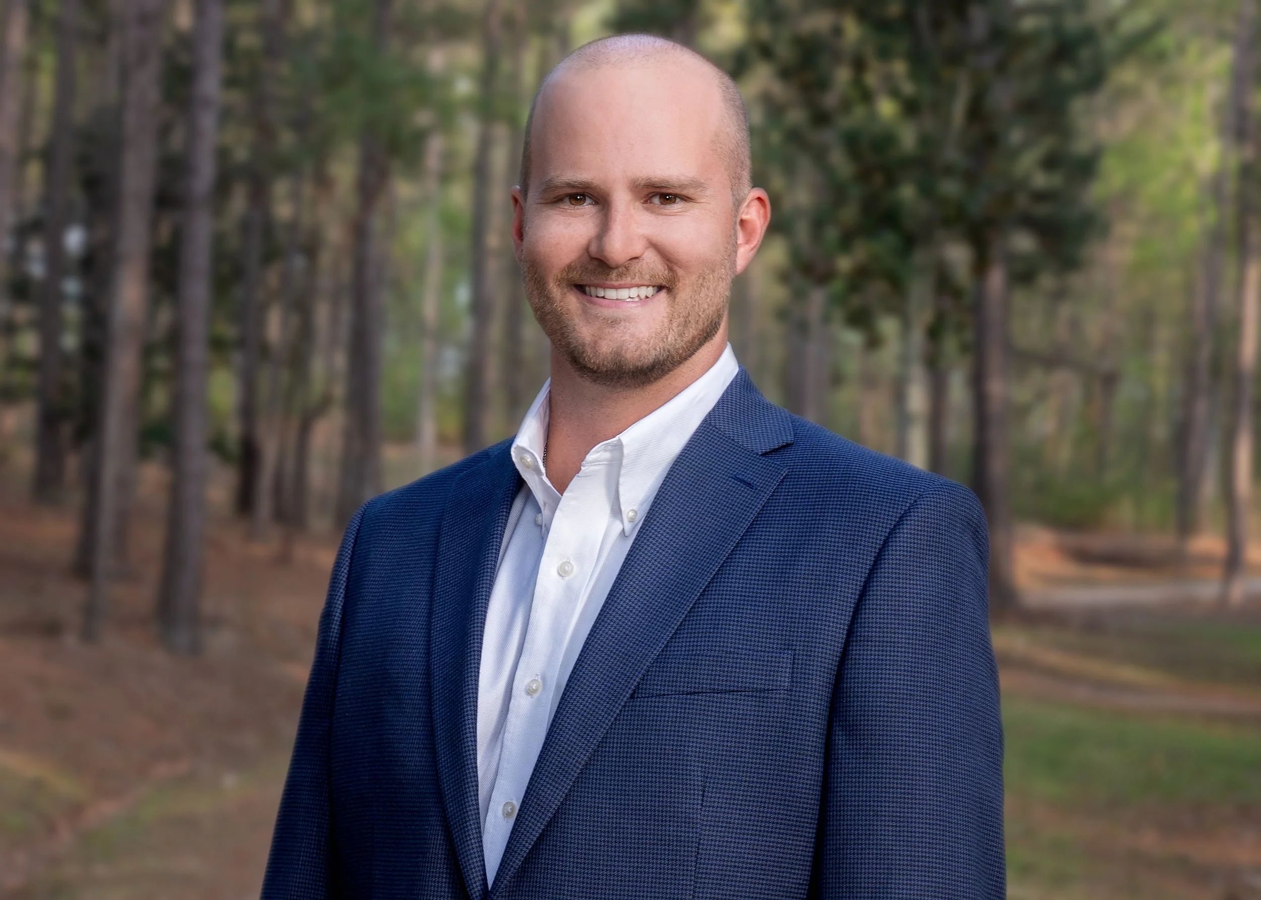 A man with a shaved head and beard, wearing a blue suit jacket and white shirt, standing outdoors in a wooded area with trees in the background.