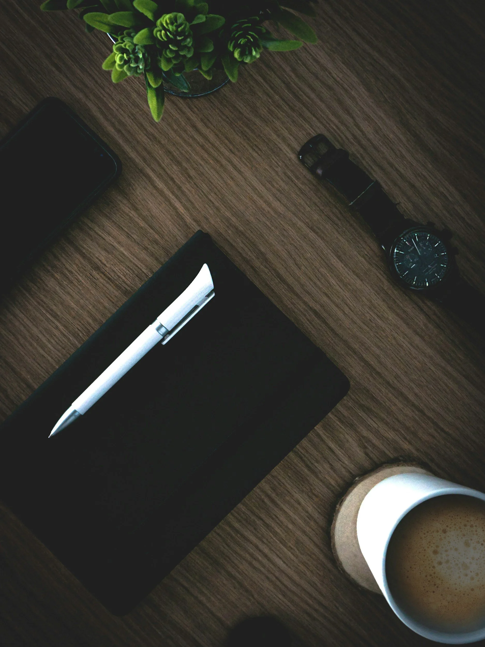 Desk setup with a coffee mug, a black notebook, a white pen, a wristwatch, a smartphone, and a small potted succulent plant on a wooden surface.