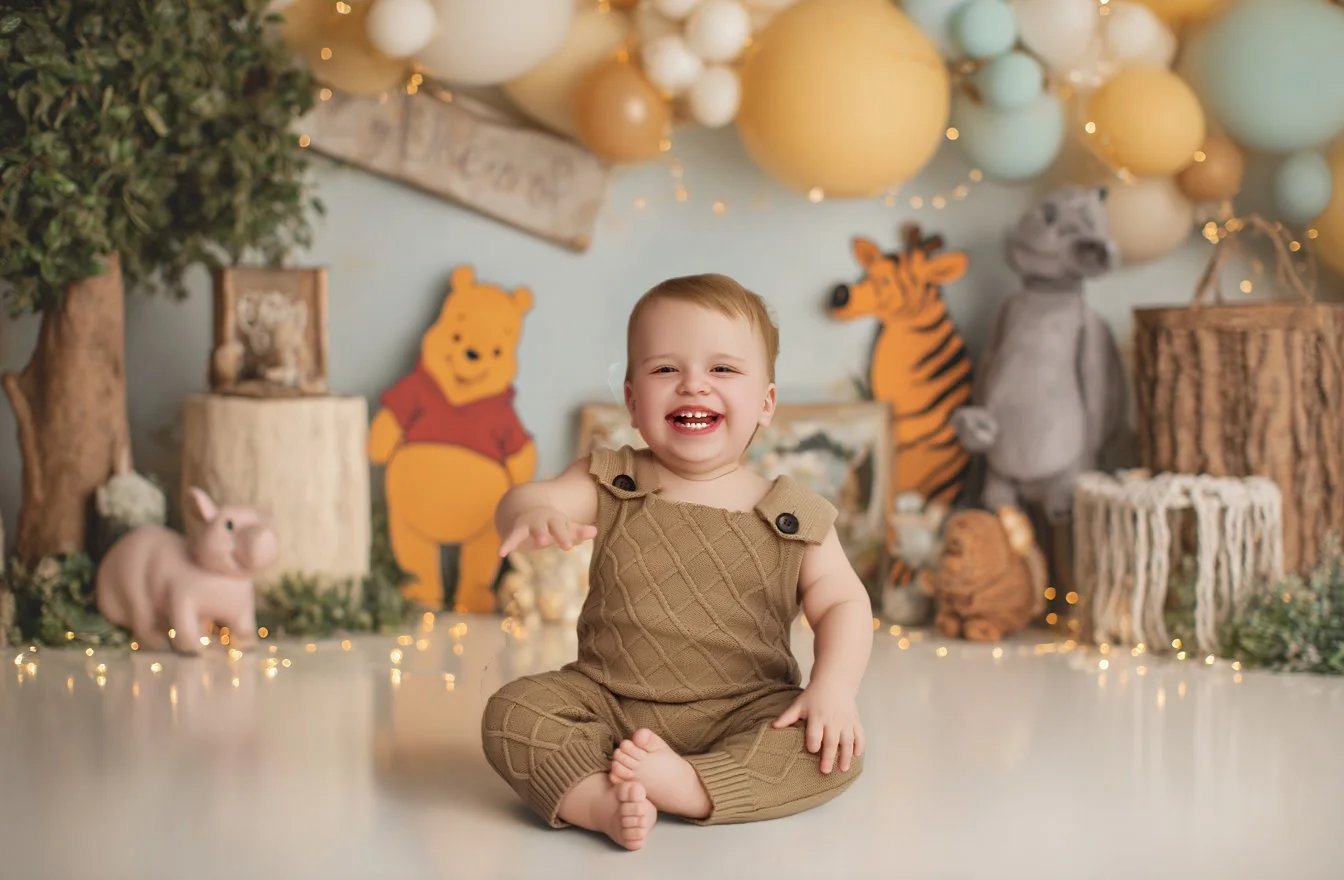 A smiling baby sitting on the floor in front of a decorated background with stuffed animal characters and balloons.