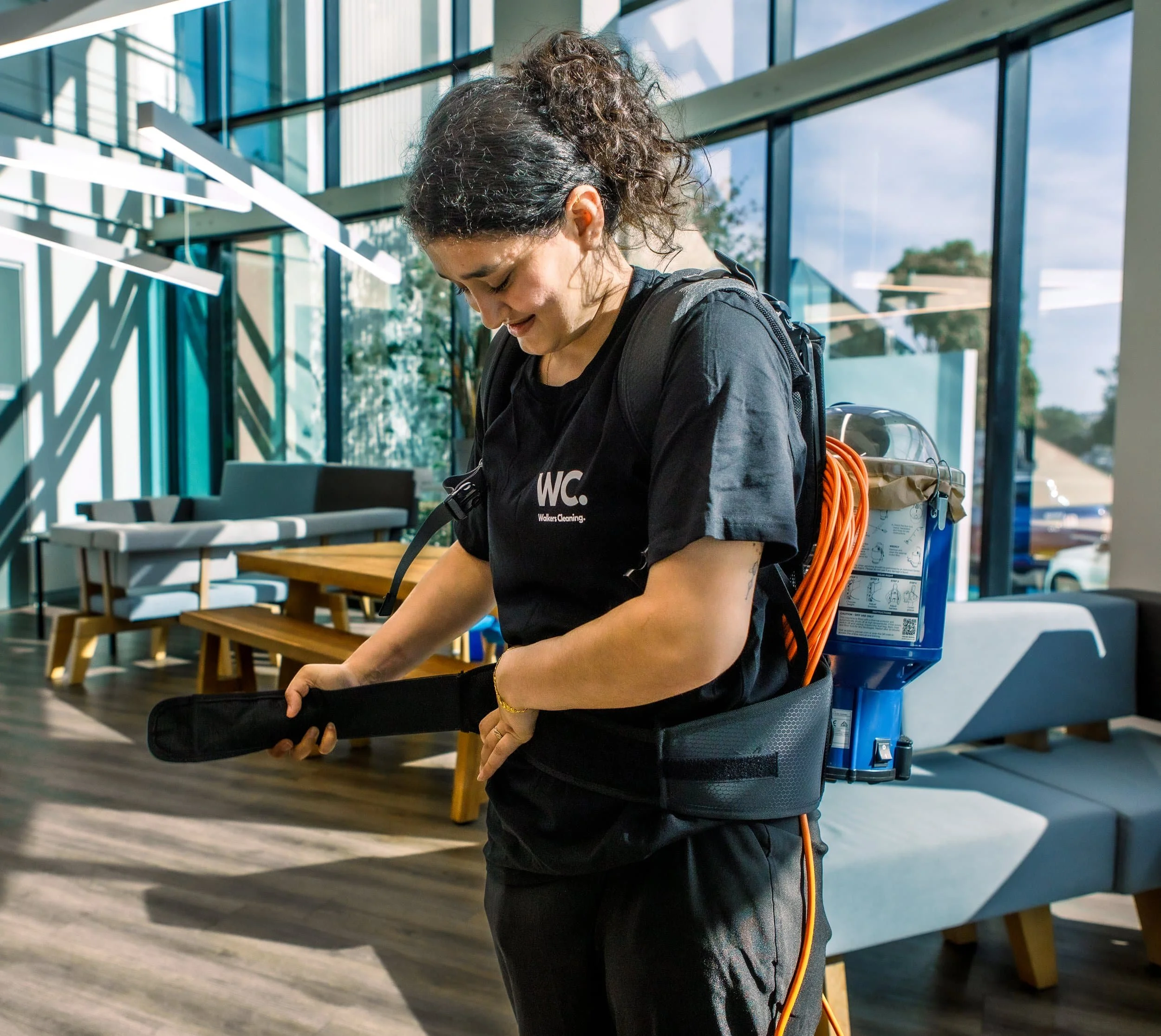 A woman in a black t-shirt and black pants is preparing cleaning equipment, standing indoors near large windows with sunlight streaming in.