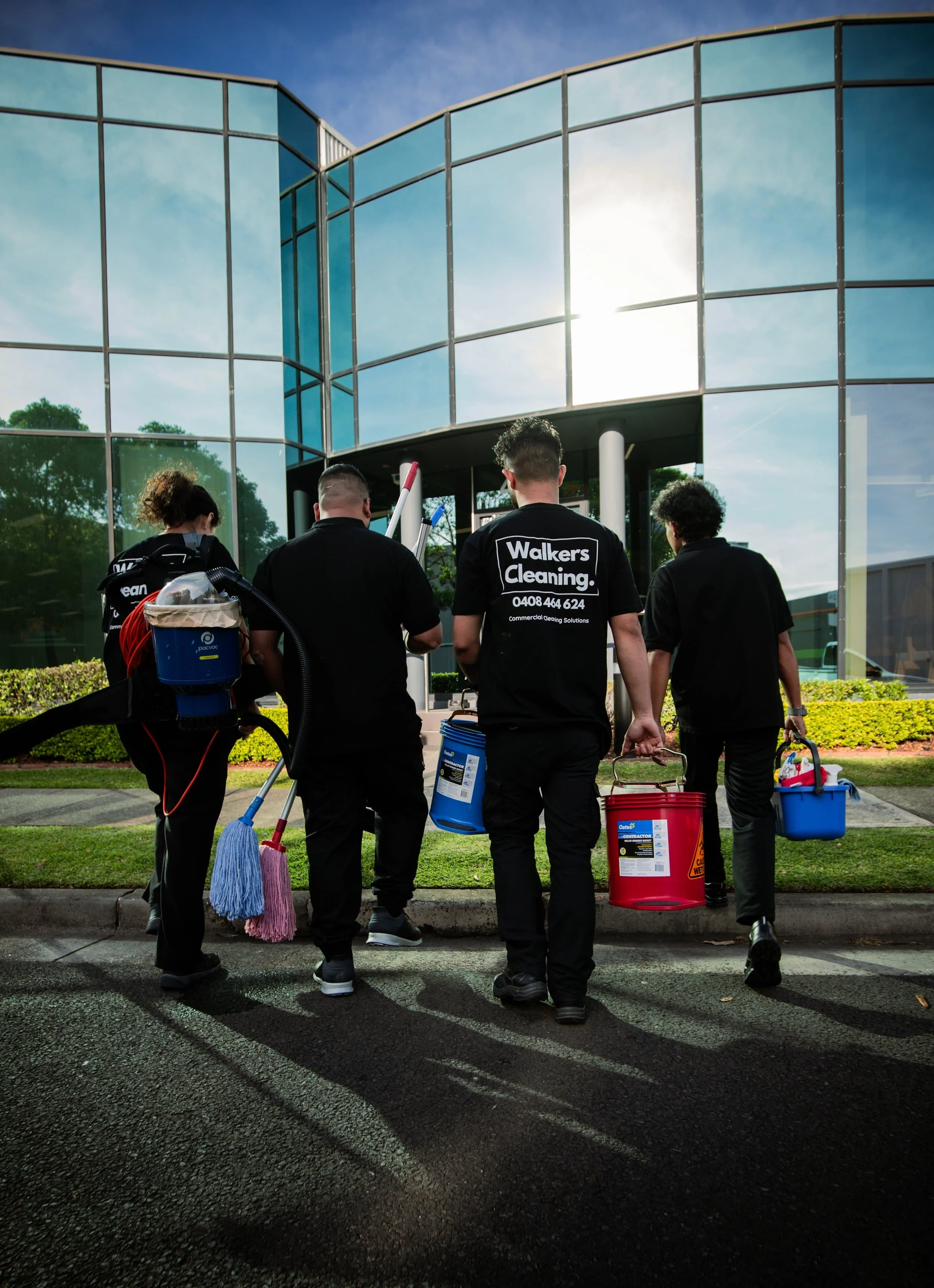 Group of professional cleaners walking toward a modern glass office building, carrying cleaning supplies and equipment on a sunny day.