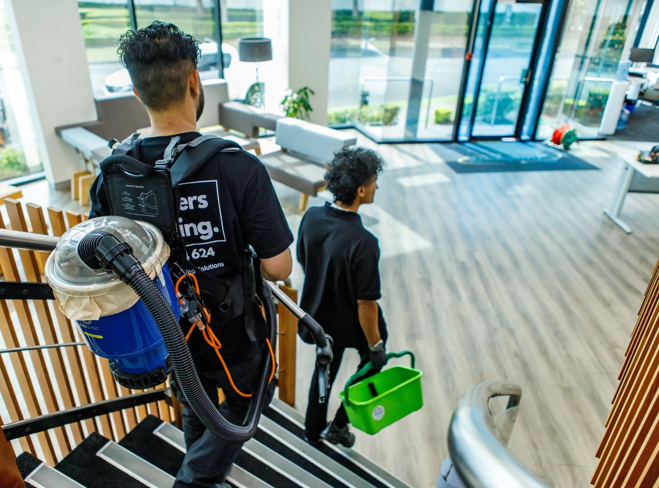 Two pest control technicians descending a staircase inside a modern building, carrying equipment and a green container.