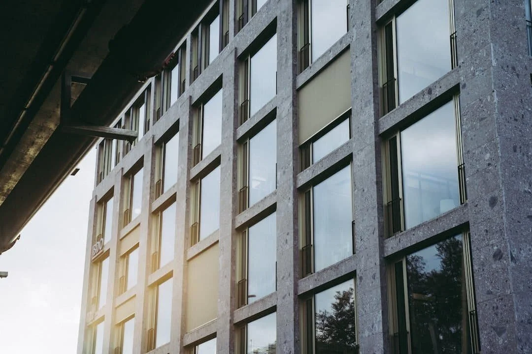 Close-up of a modern multi-story building with large reflective windows and stone facade, with sunlight creating a glow on the left side.