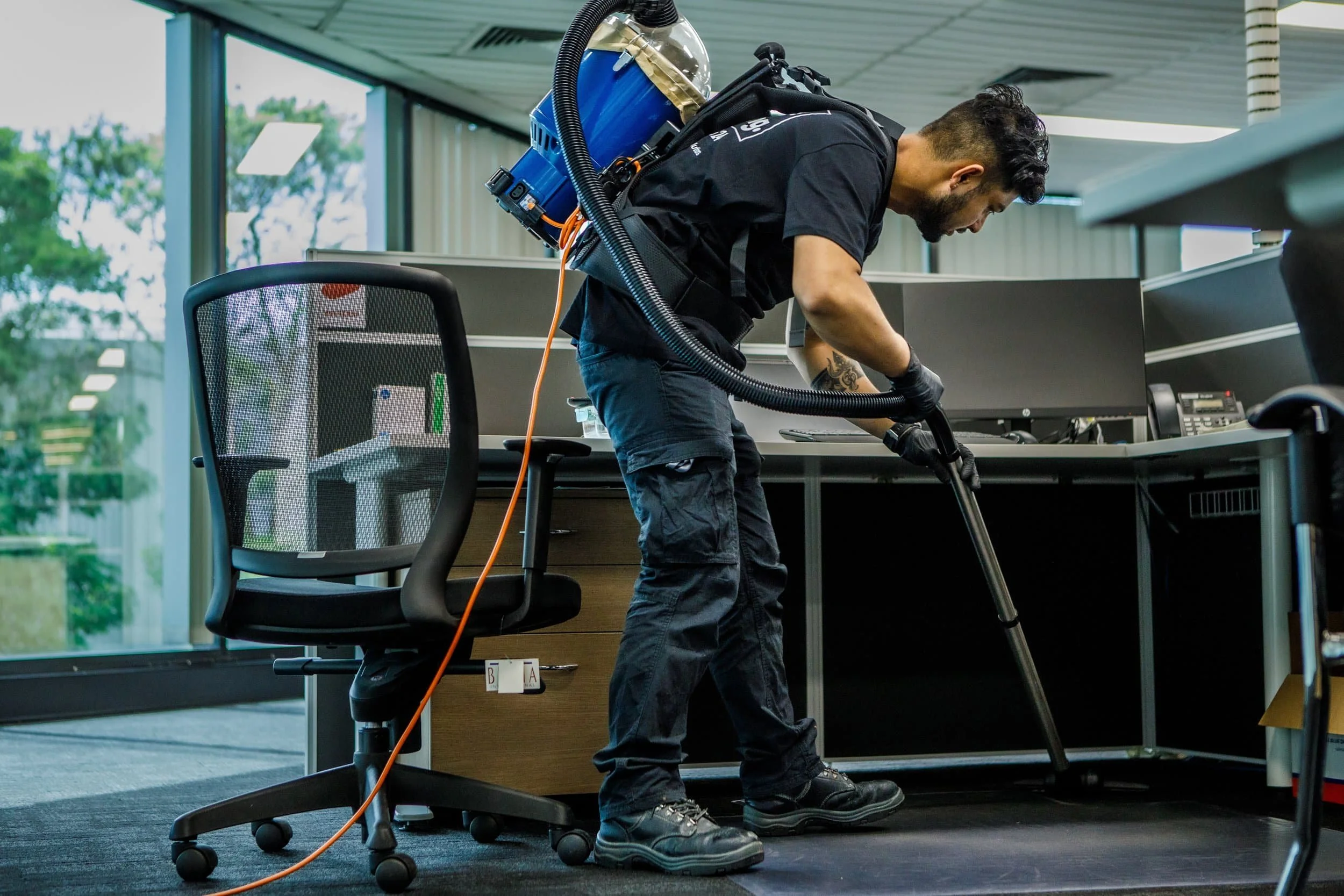 A man in black clothes and gloves cleaning an office floor with a professional steam cleaning machine. The office has large windows, an office chair, and desks with computer monitors.