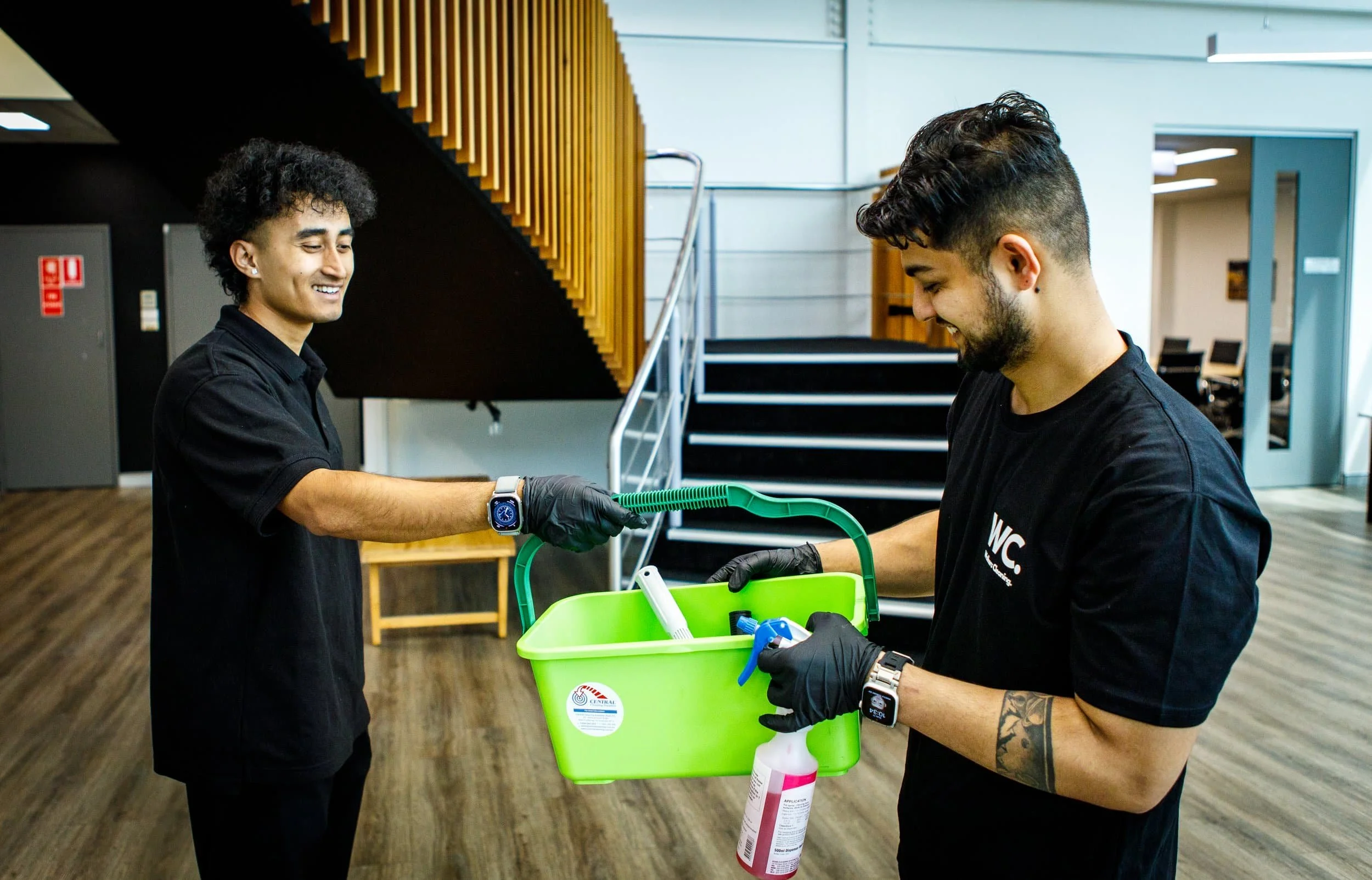 Two men wearing black shirts and black gloves exchanging cleaning supplies with a green bucket in a modern office space.
