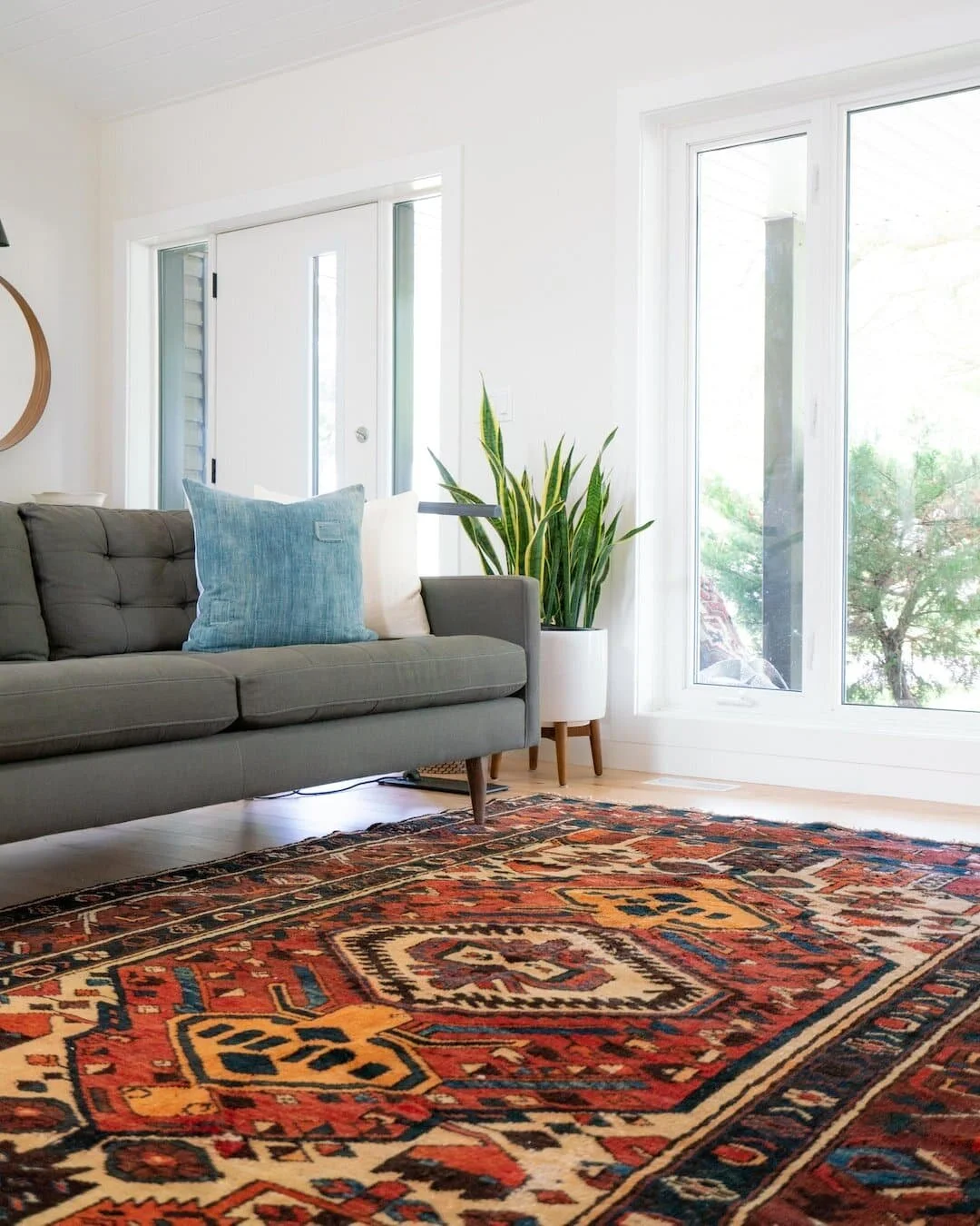 Living room with a grey sofa, blue and white cushions, potted plant, large window, and a colorful patterned rug.