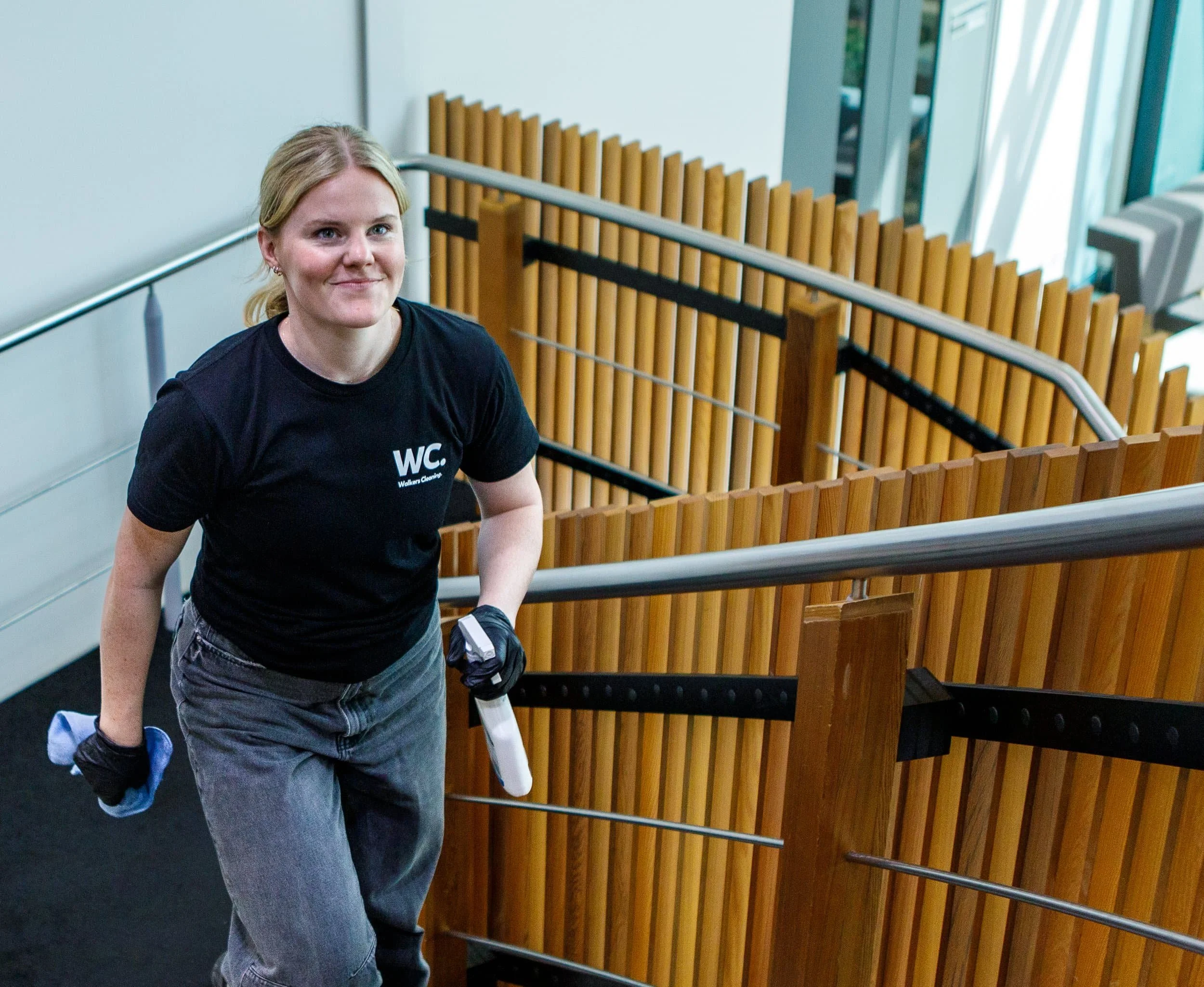 A person with blonde hair wearing a black t-shirt and gray pants, holding cleaning tools, ascending a stairs in a modern indoor setting with wooden paneling and large windows.