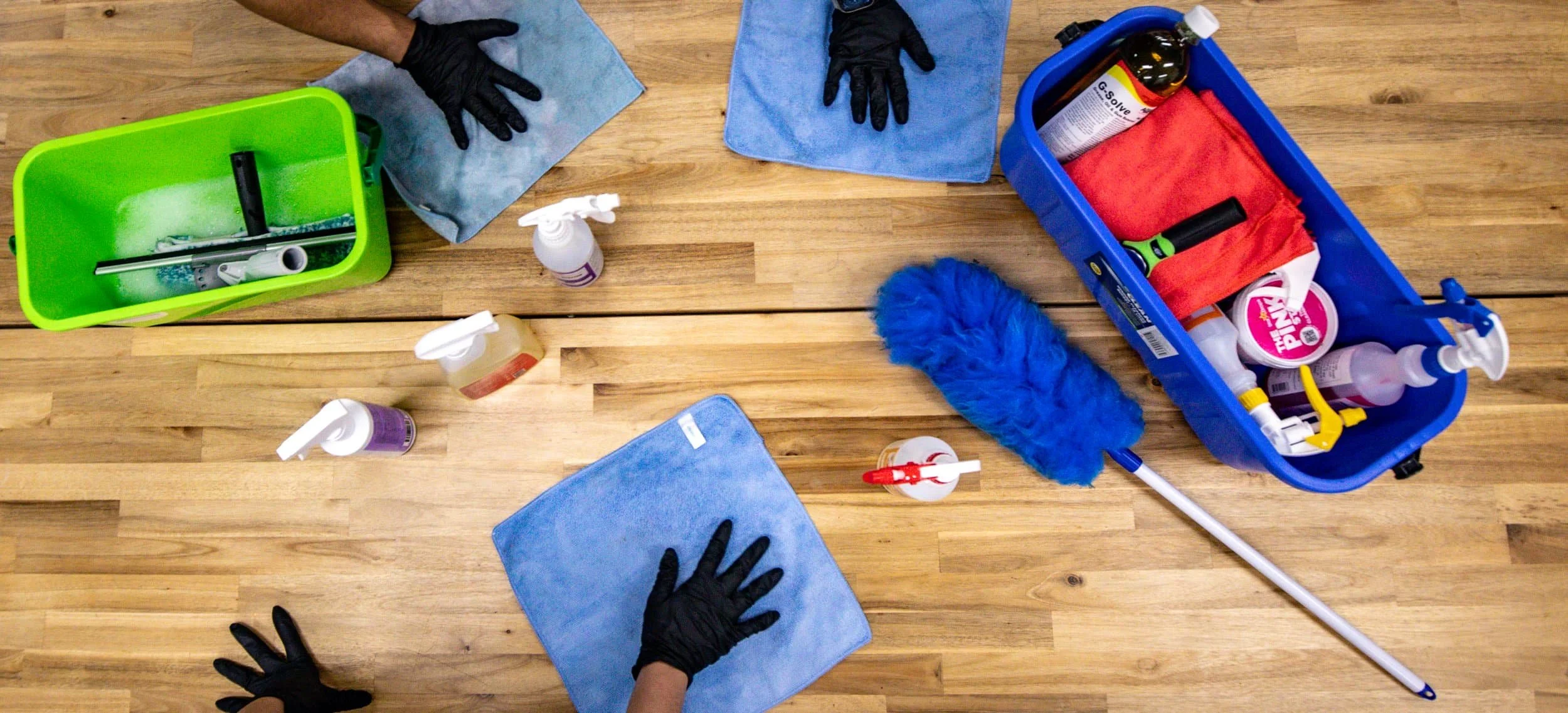 Cleaning supplies on a wooden floor, including spray bottles, microfiber cloths, a blue duster, a mop, and gloves.