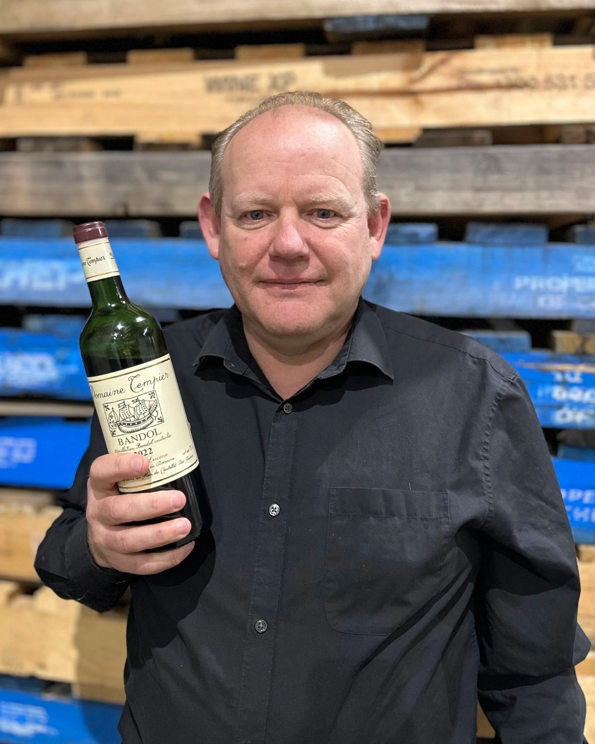 A man wearing a black shirt holding a bottle of red wine, standing in front of wooden pallets.