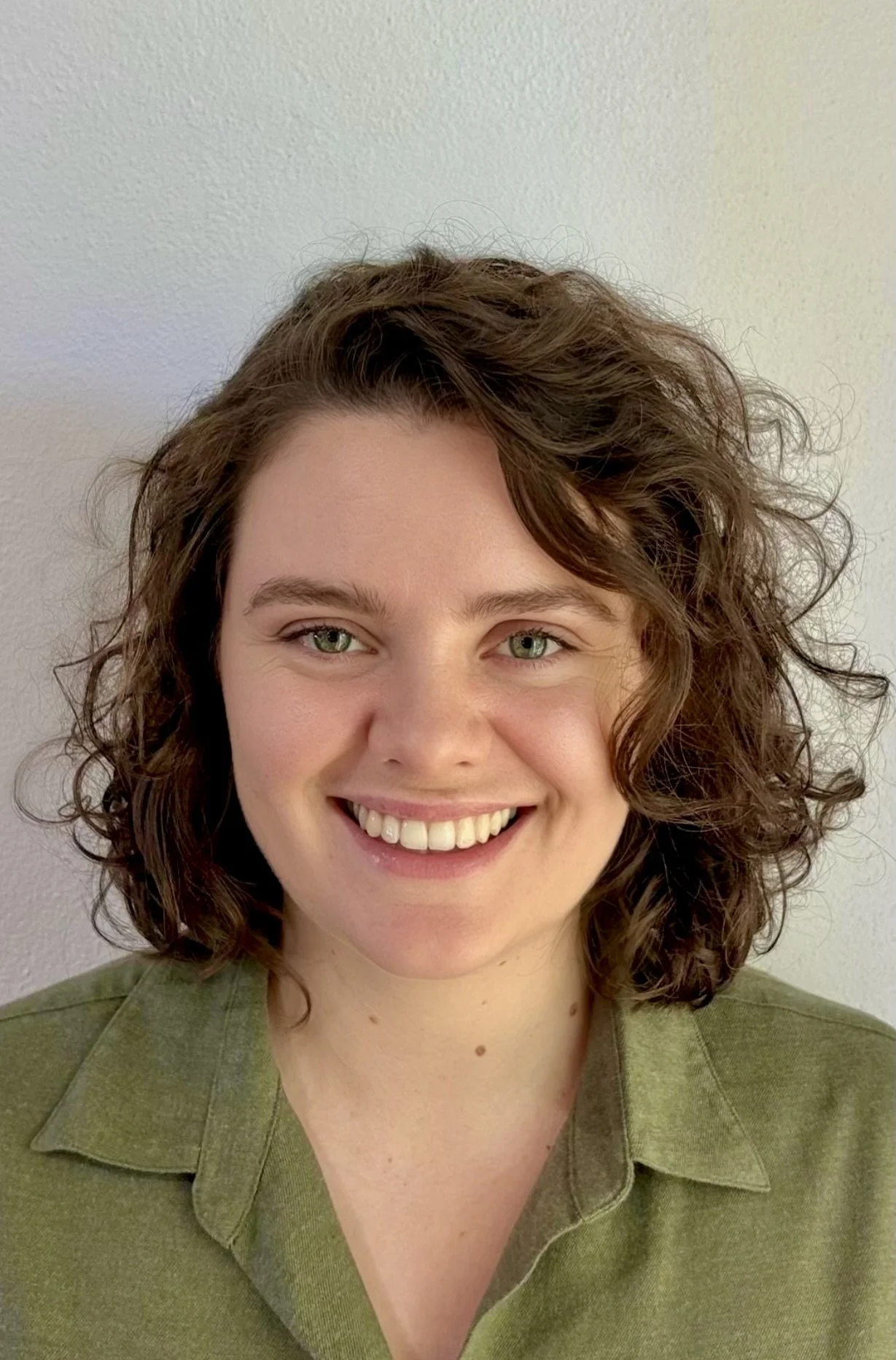 A woman smiling with curly brown hair, green eyes, and wearing an olive green shirt, standing against a plain light-colored wall.
