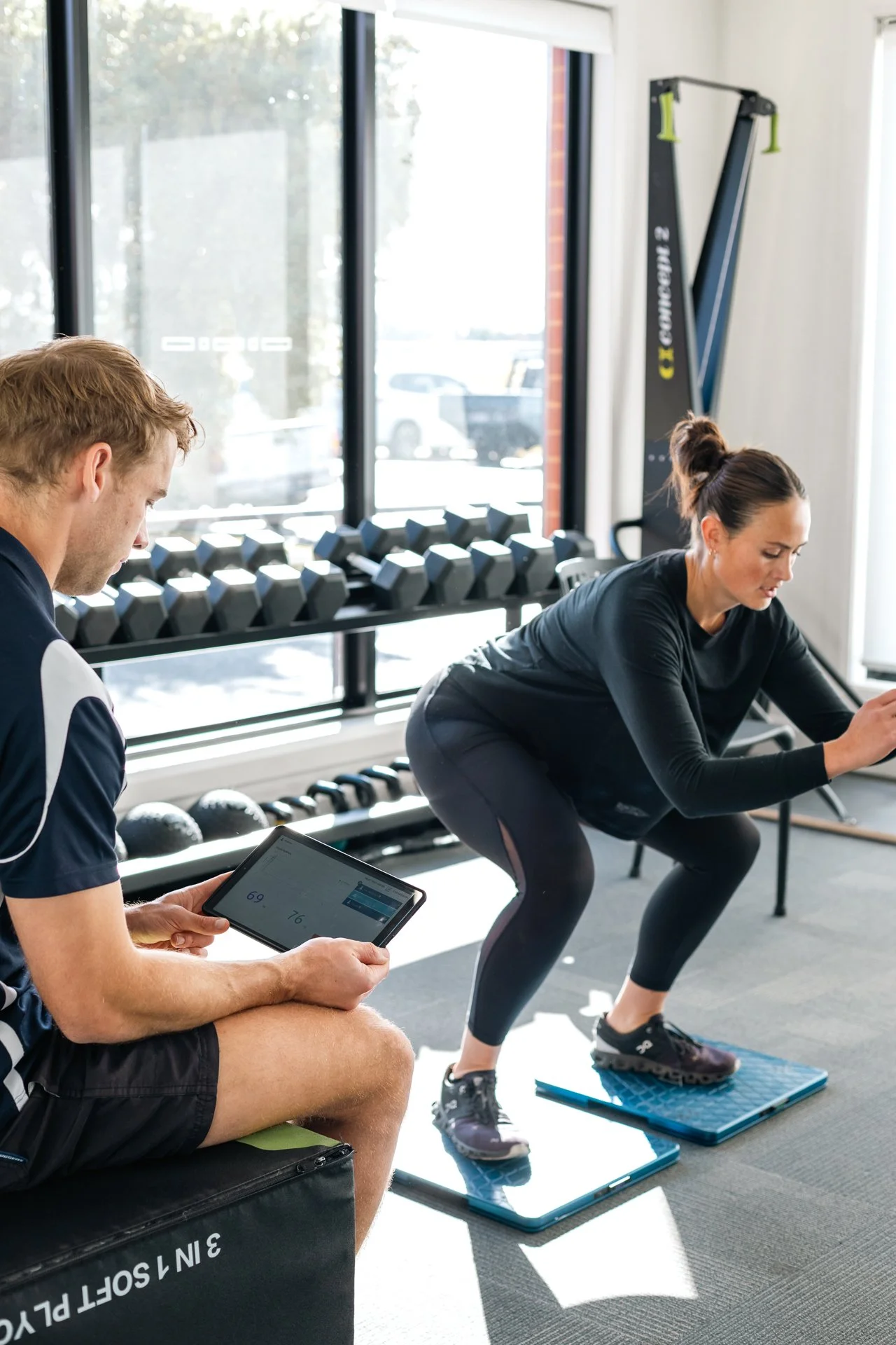 Woman in black workout clothes doing squats on digital scale while man in shorts and sports shirt sitting on a bench taking notes on a tablet in a gym.