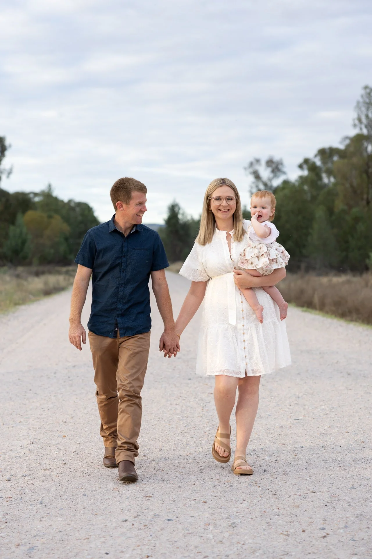 A family of three walking hand in hand on a gravel road outdoors during daytime, with trees and cloudy sky in the background. The woman is carrying a young child and smiling, while the man is looking at her and smiling.