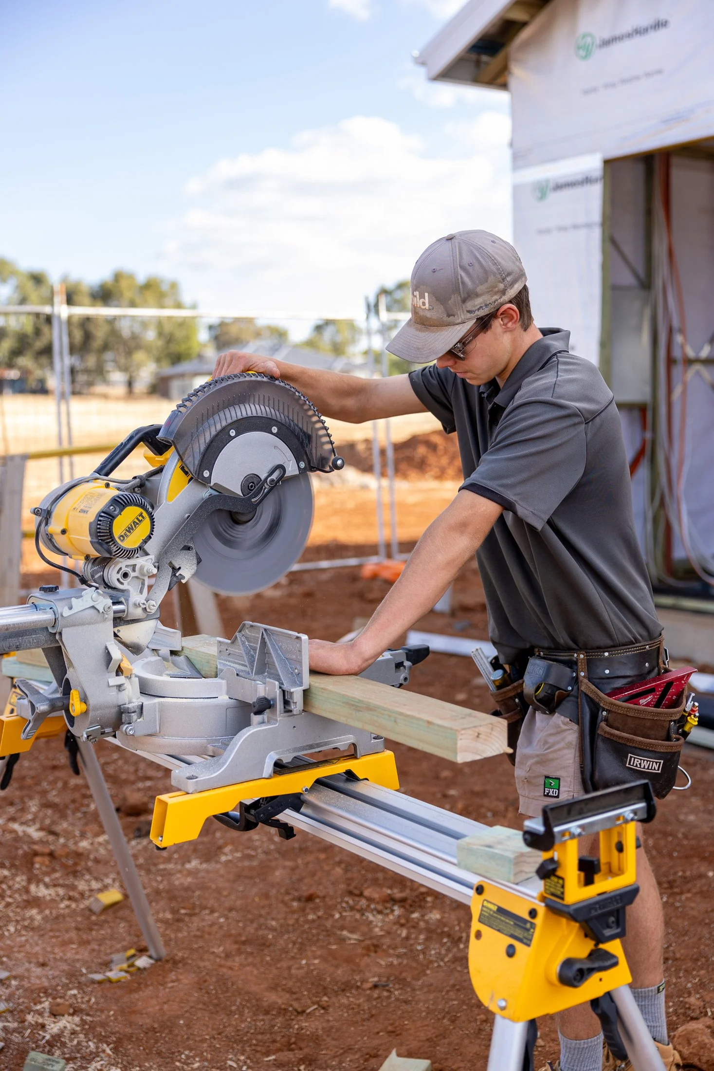 A young man working on a construction site using a miter saw to cut a piece of wood. He is wearing a gray cap, glasses, a dark gray shirt, and work pants with tools in his pocket. The background shows open land and a partially built structure.