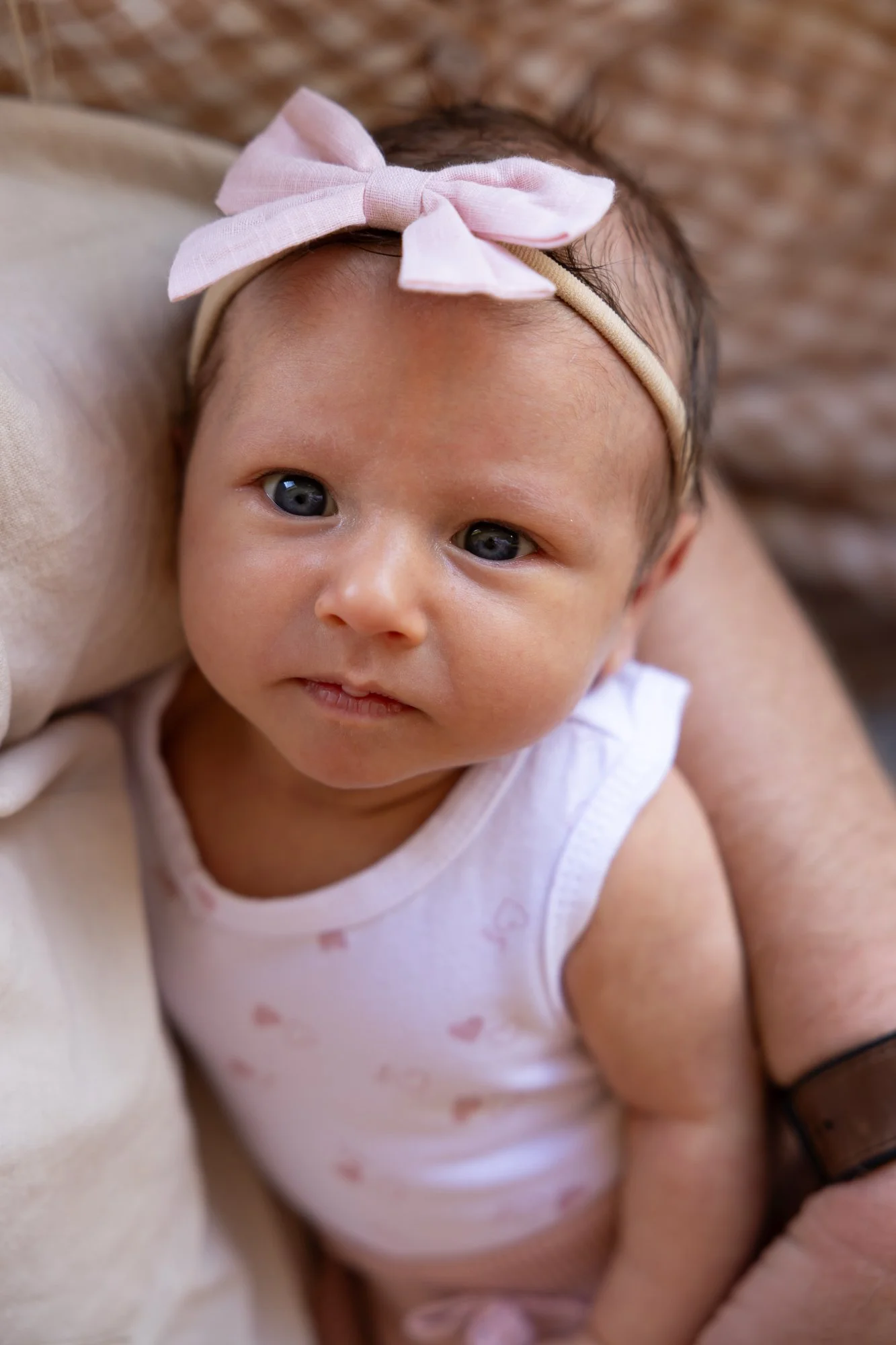 Close-up of a baby girl with dark hair, wearing a beige headband with a pink bow and a white sleeveless top with pink heart patterns, resting on someone's arm.