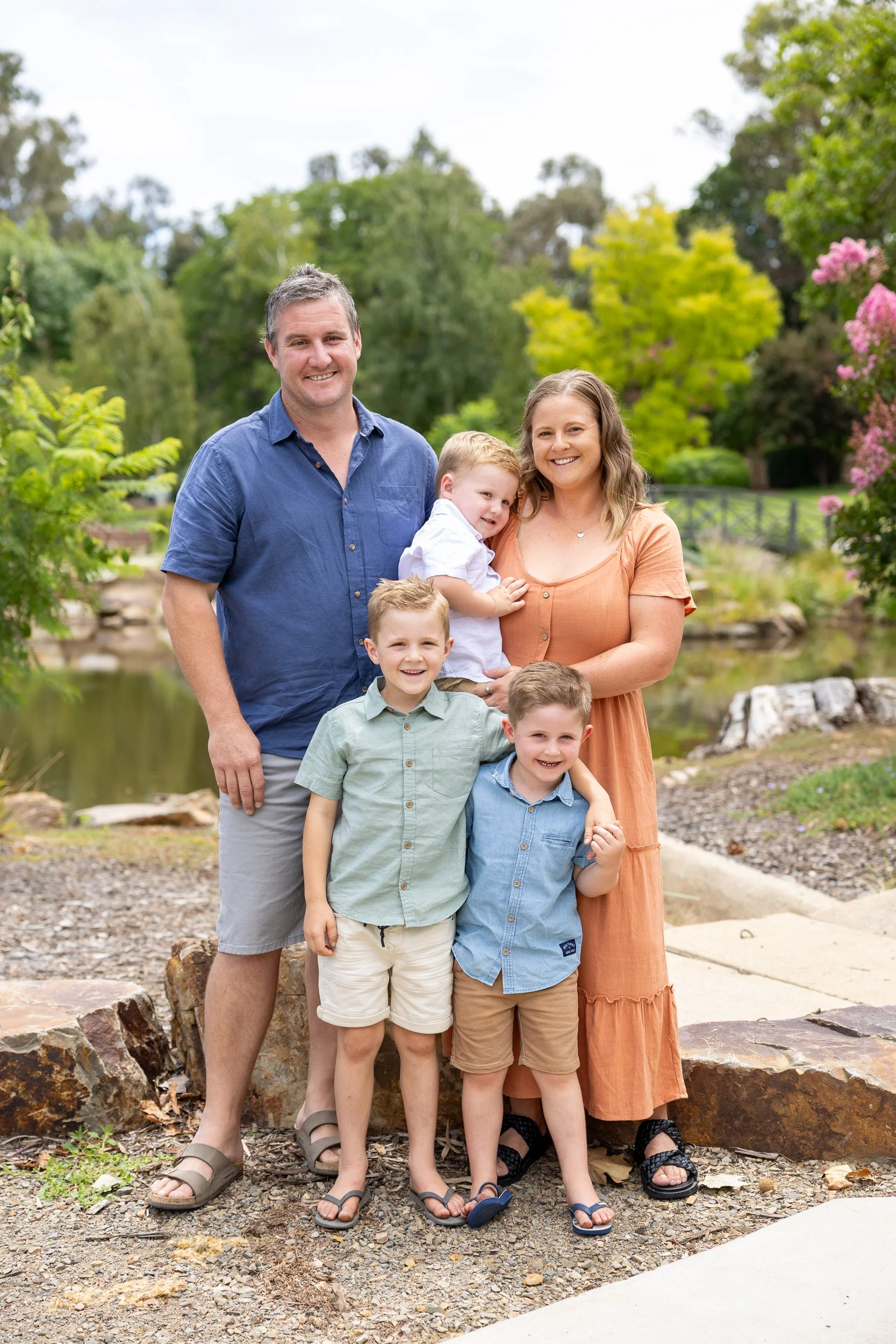 A family of six standing outdoors near a pond, surrounded by trees and greenery.