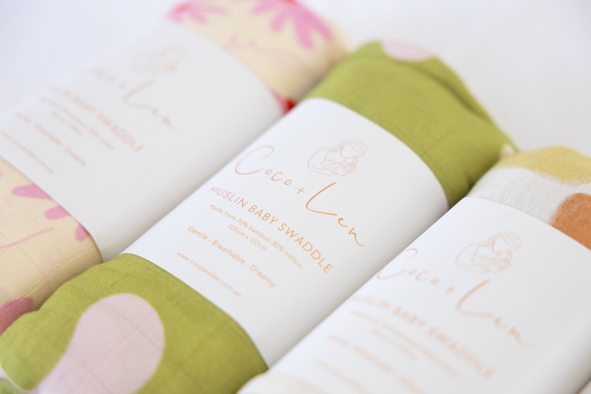 Close-up of eco-friendly baby swaddles in floral and pastel green fabric, wrapped in white labels with the brand name 'Coco + Lune' and product description.