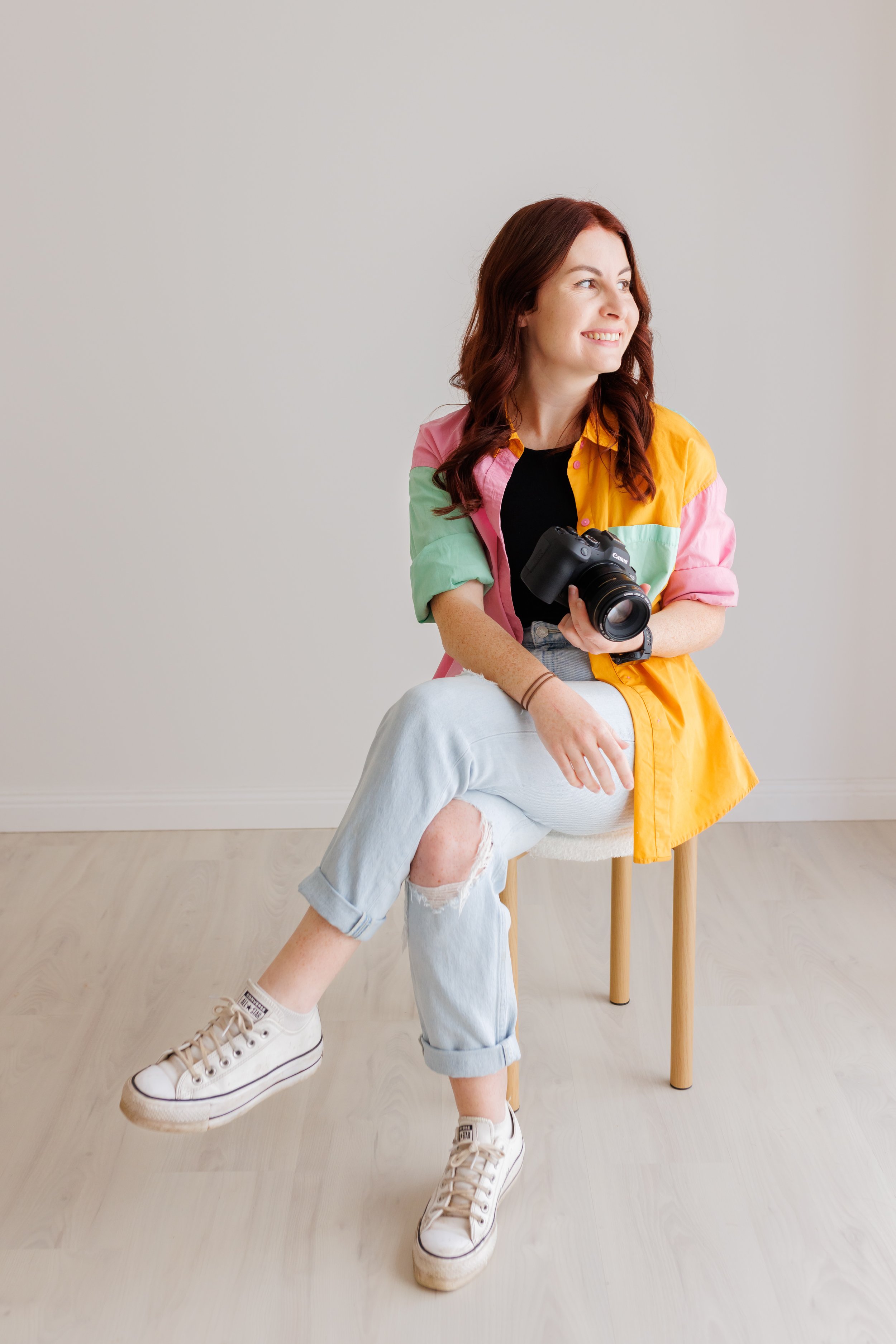 A young woman with red hair in a colorful shirt and jeans sitting on a stool, holding a camera, smiling and looking to her right indoors.