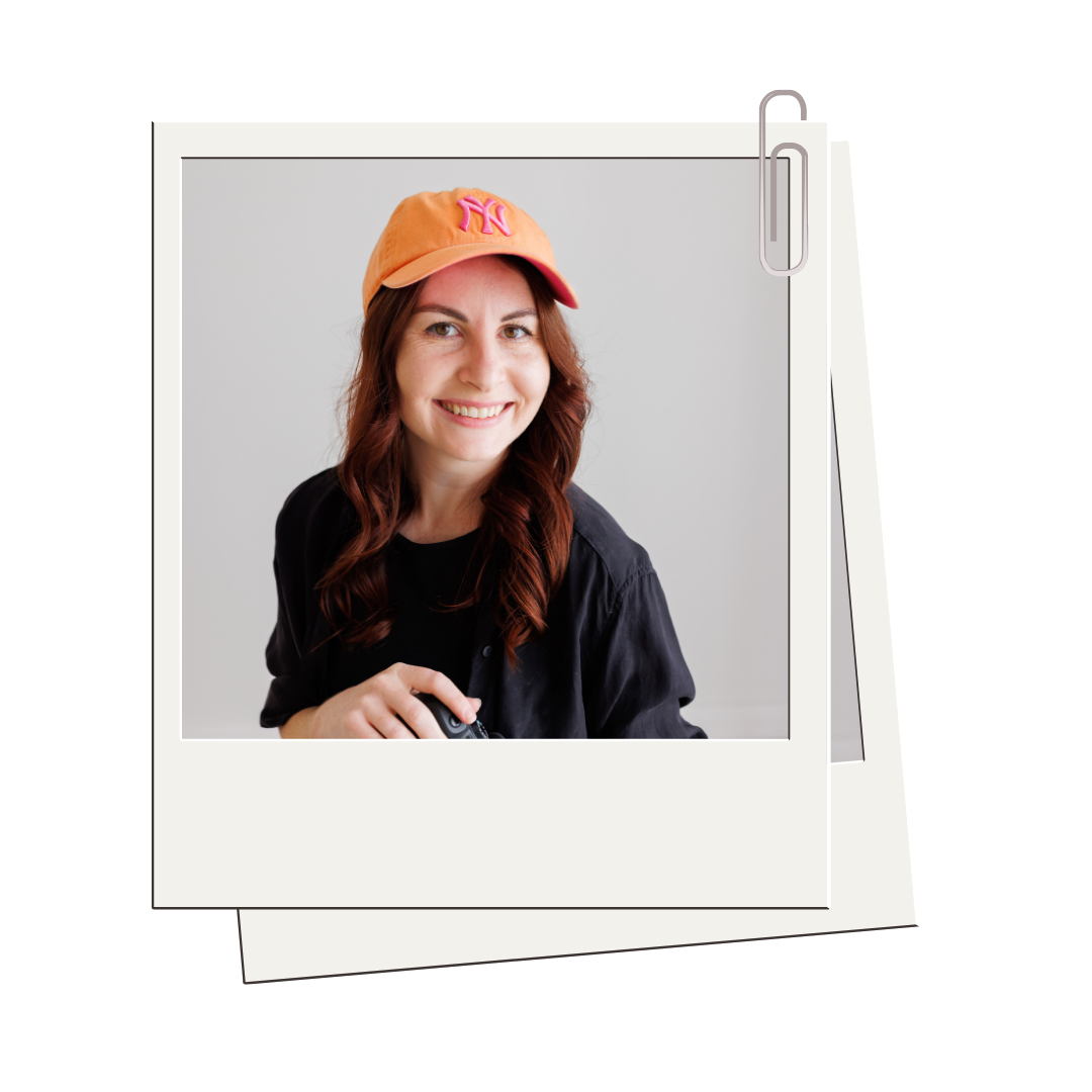 Photograph of a smiling woman wearing a pink and orange baseball cap, with long wavy hair, in a casual black shirt, holding a camera, against a plain gray background.