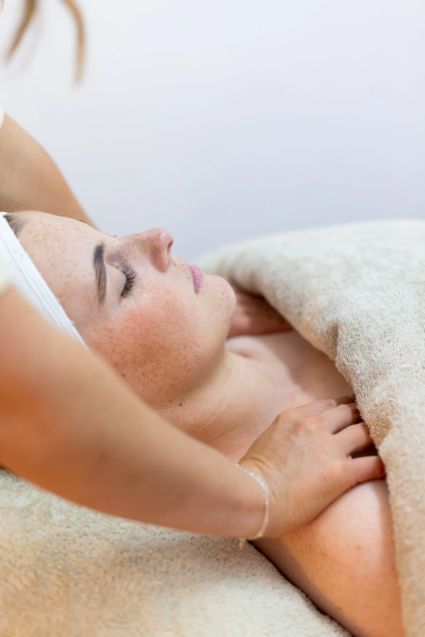 A woman receiving a massage, lying on a massage table with a towel over her, eyes closed, in a spa or wellness center.