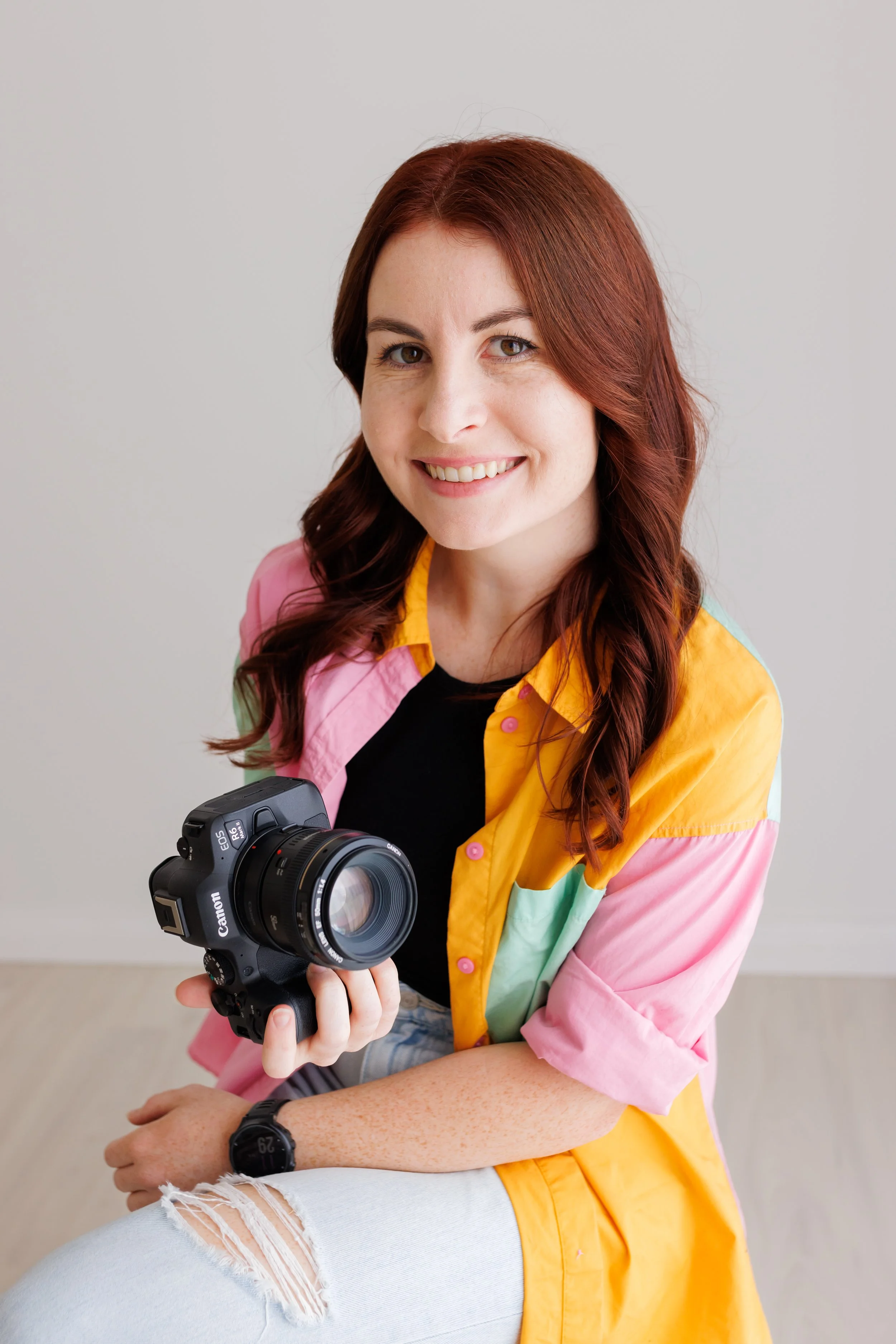 A woman with red hair holding a Canon camera, sitting against a plain light-colored wall.