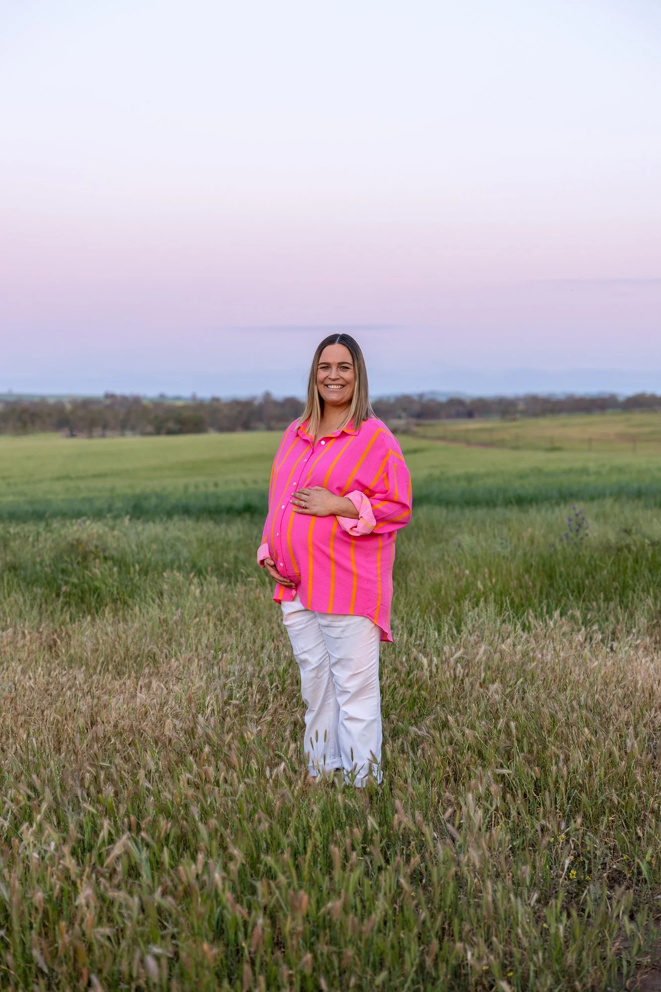 A pregnant woman standing in a grassy field at sunset, smiling and wearing a pink and yellow striped shirt with white pants.