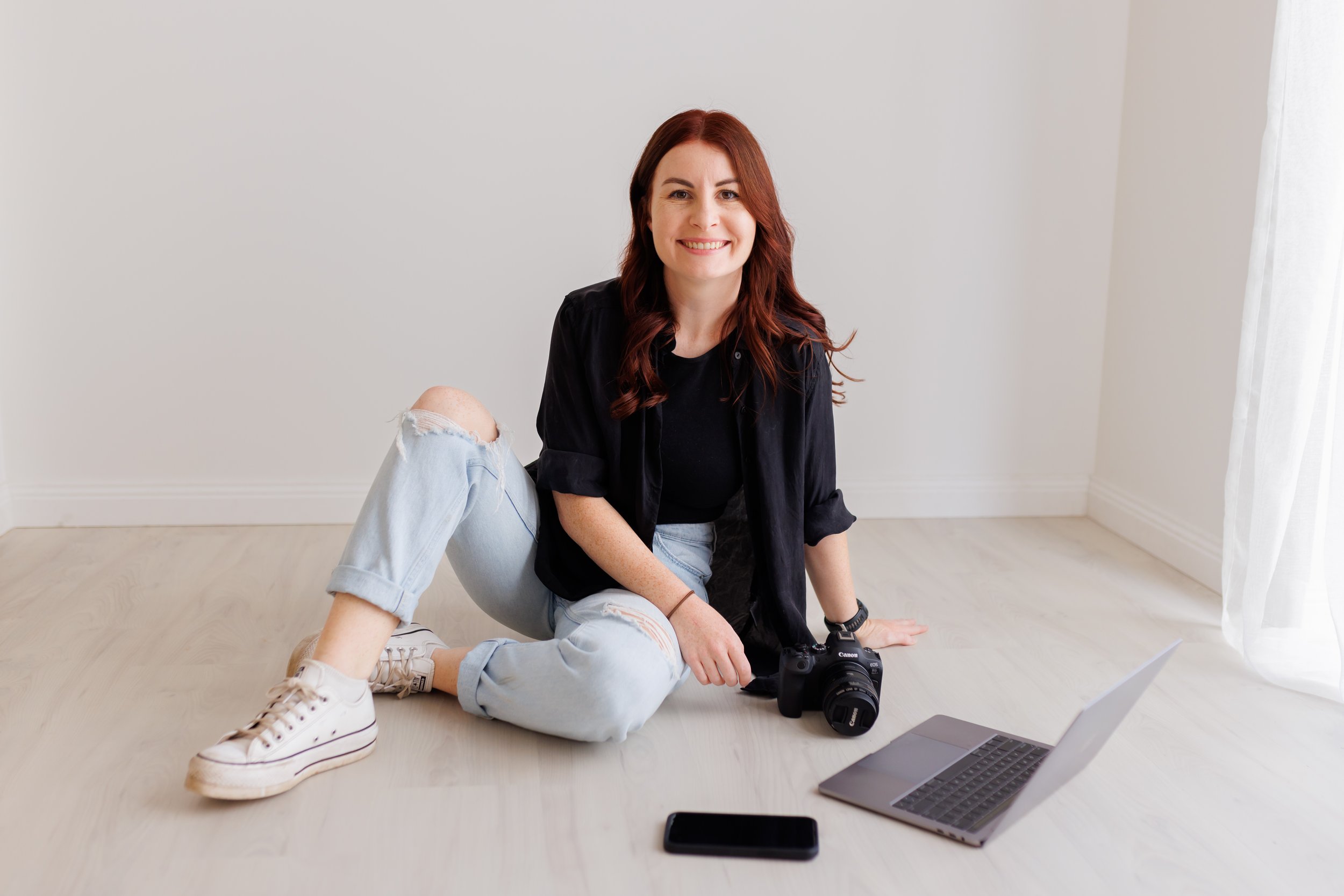 Young woman with red hair sitting on the floor, smiling, with a camera, laptop, and smartphone in a bright room with white walls and light-colored floor.