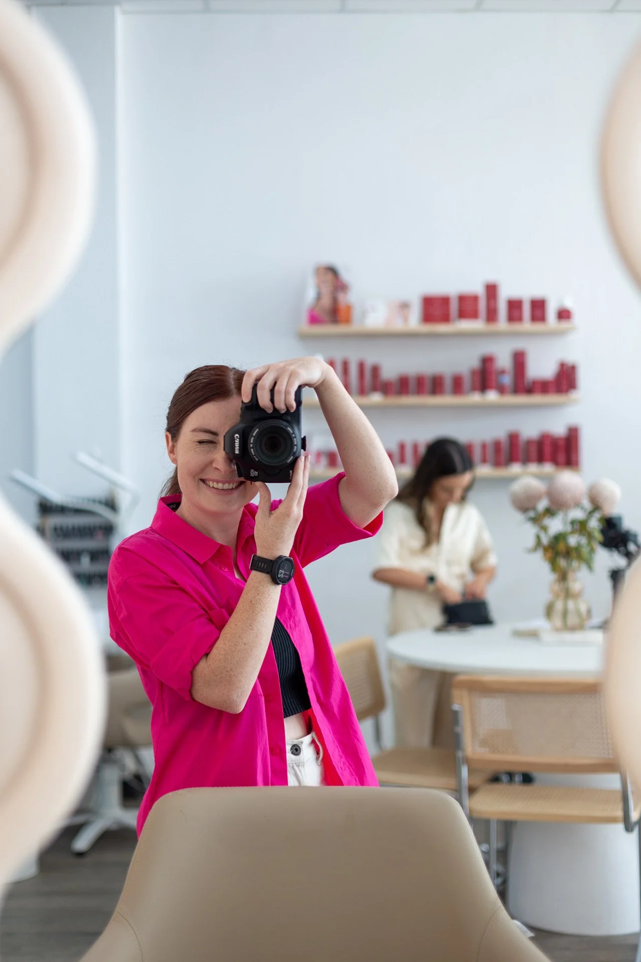 A woman with brown hair, wearing a bright pink shirt, taking a selfie with a camera while smiling. In the background, another woman is looking at her phone, with shelves of red-colored products and a vase of pink flowers on a white table.