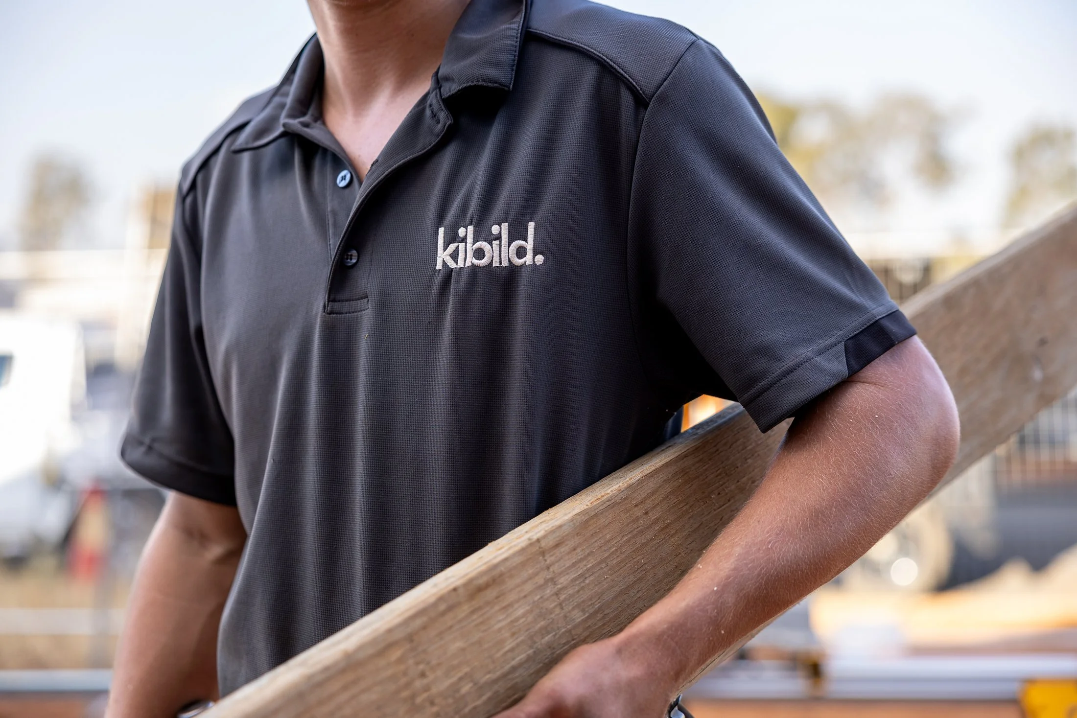 Close-up of a person wearing a black polo shirt with the logo 'kibild' on it, holding a piece of wood at a construction site.