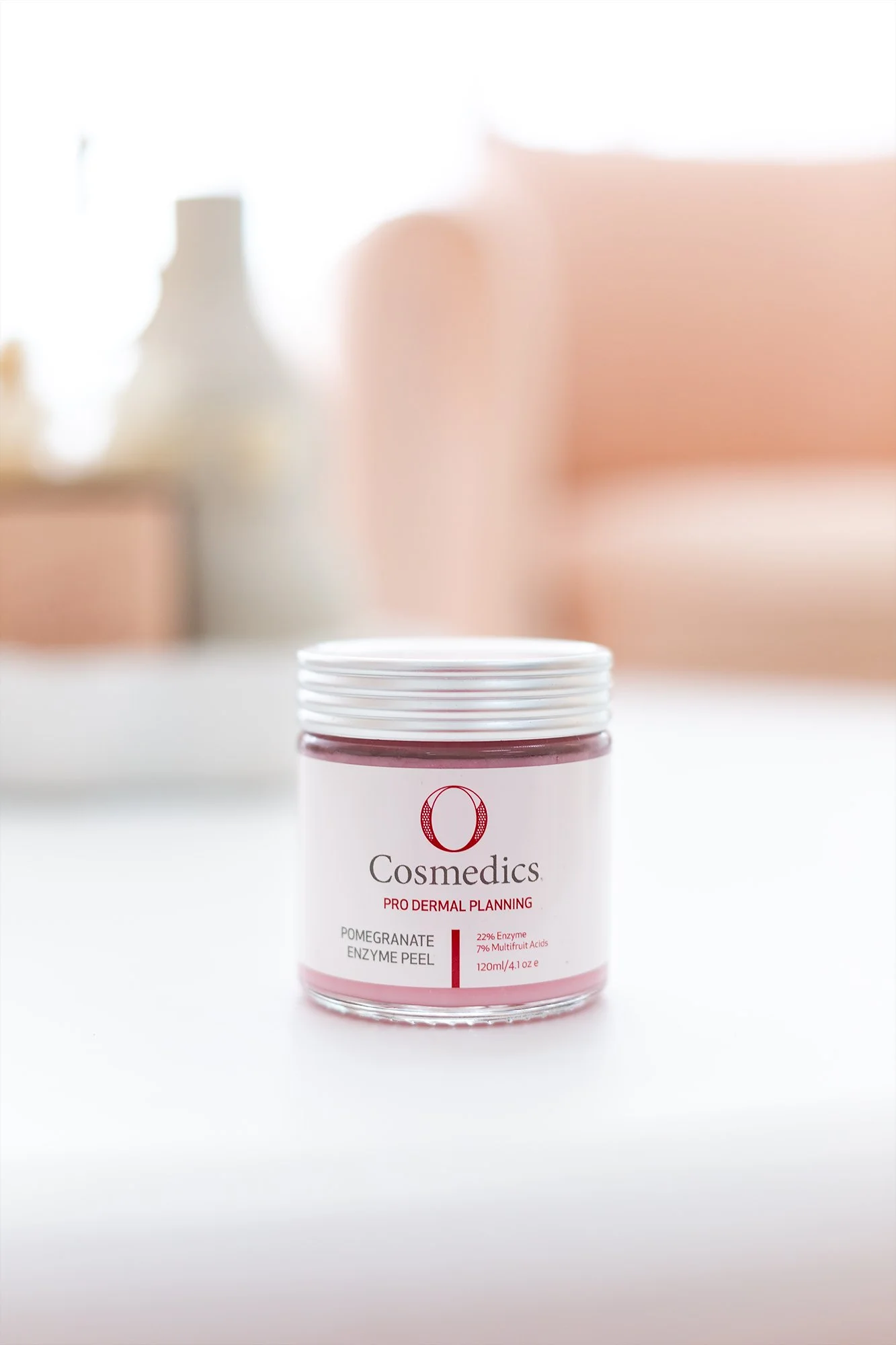 A jar of Cosmedics Pomegranate Enzyme Peel on a white surface with a blurred background of a pink sofa and white decor.