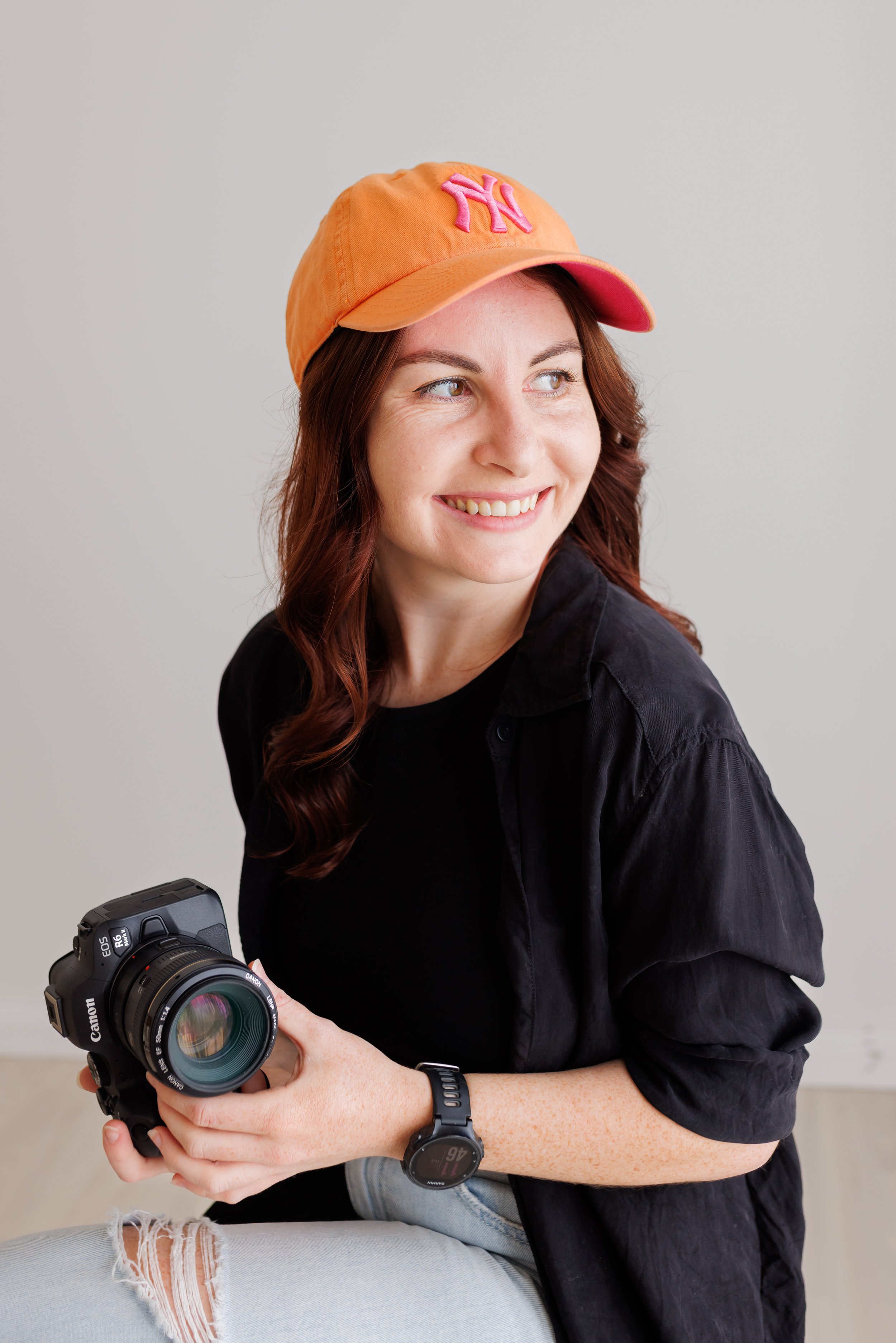 A woman with red hair wearing an orange baseball cap, black shirt, light blue ripped jeans, and a fitness tracker, holding a Canon EOS camera and smiling while looking to her right.