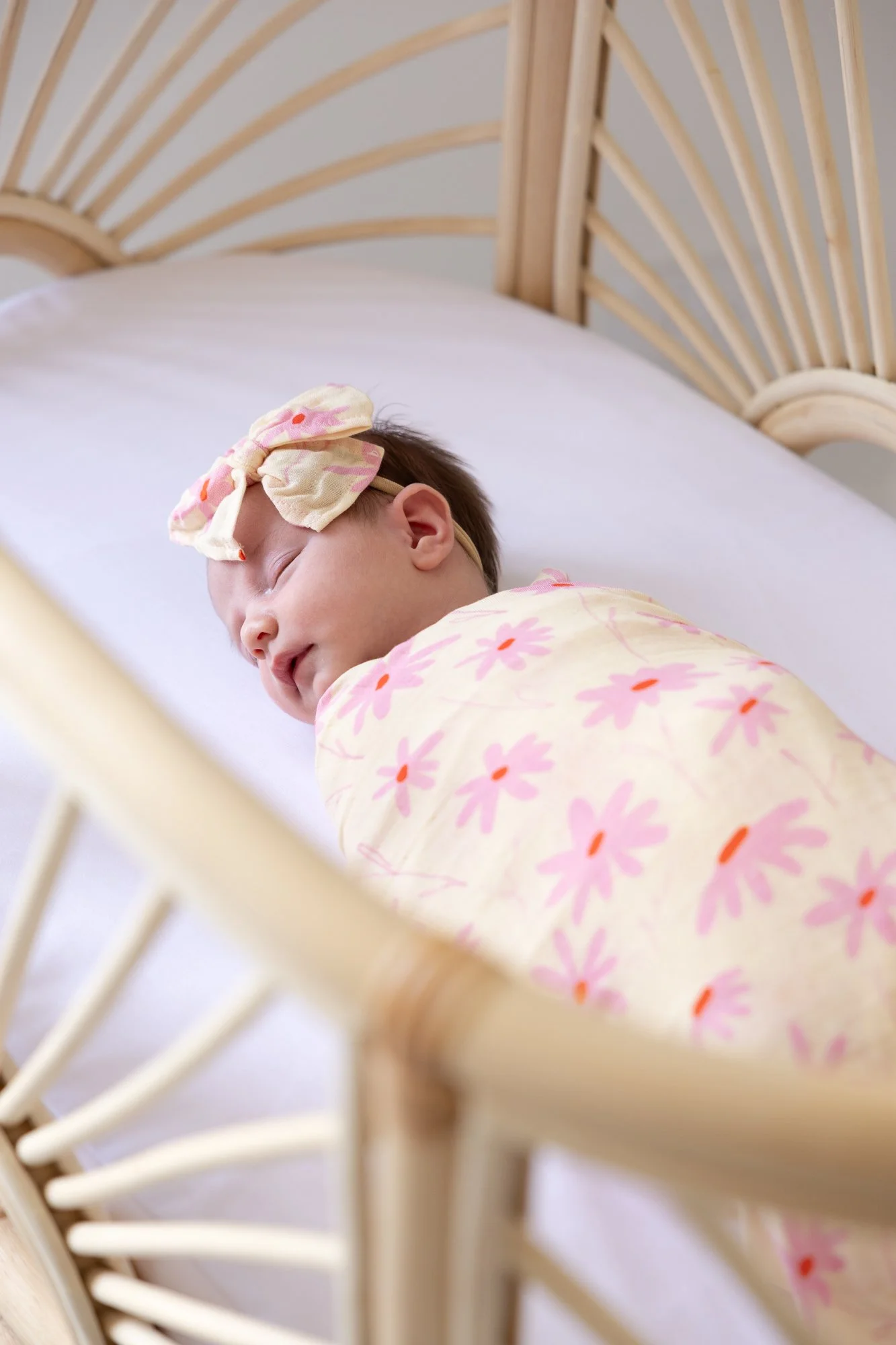 A young girl sleeping in a bed with a rattan headboard, wrapped in a floral blanket and wearing a matching bow headband.