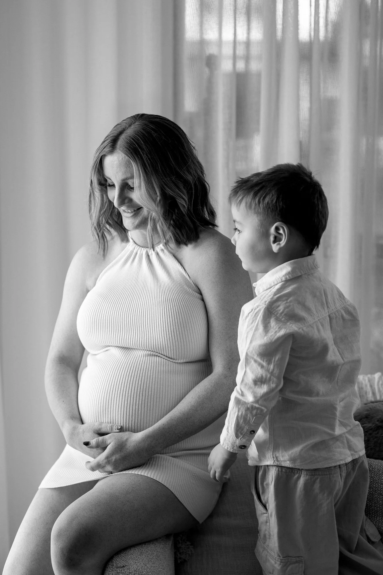 A pregnant woman sitting on a sofa with an older boy standing beside her, both smiling, near a window with curtains in background, black and white photo.