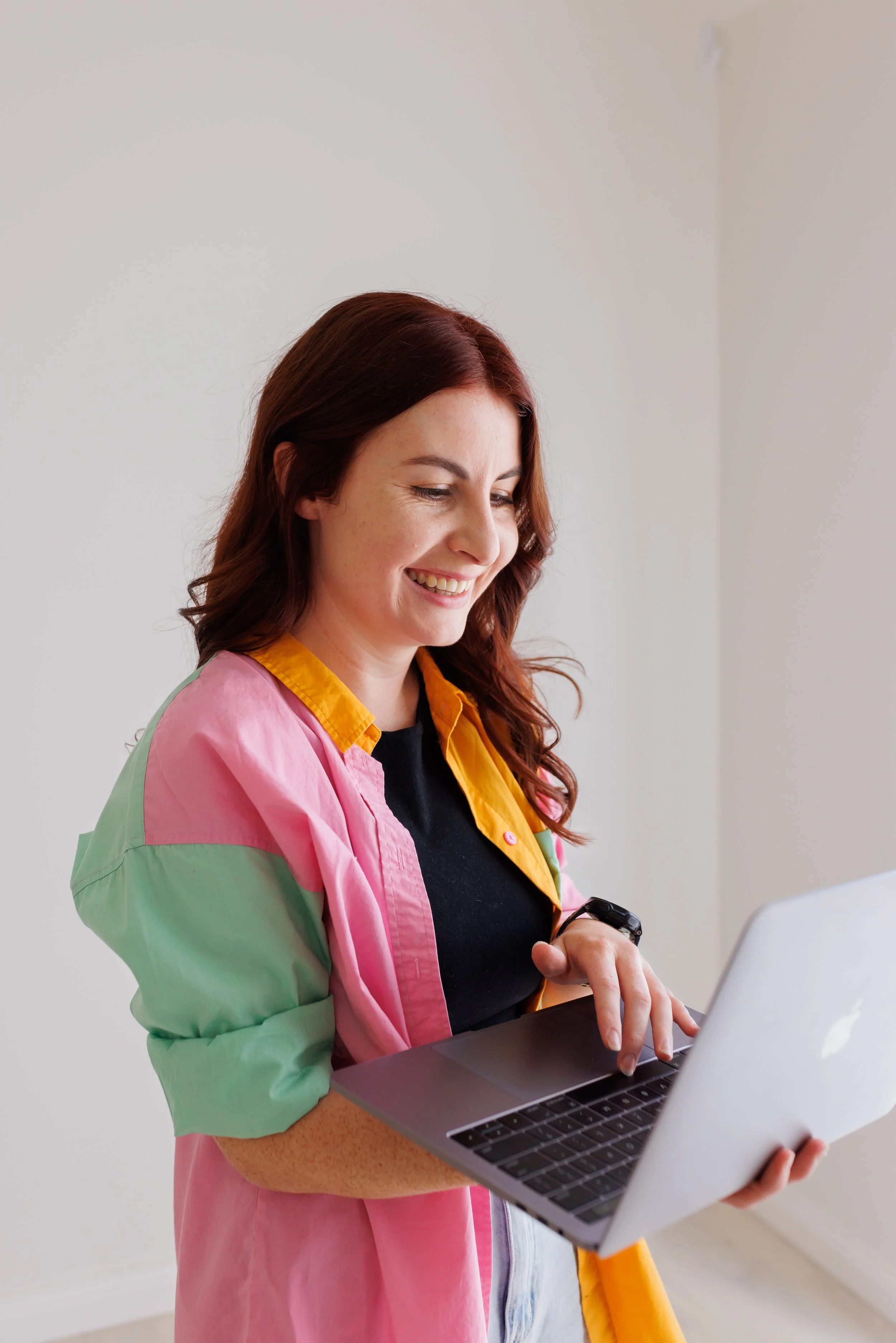 A woman with red hair smiling and using a silver laptop, wearing a colorful pink, green, and yellow jacket and a black shirt.