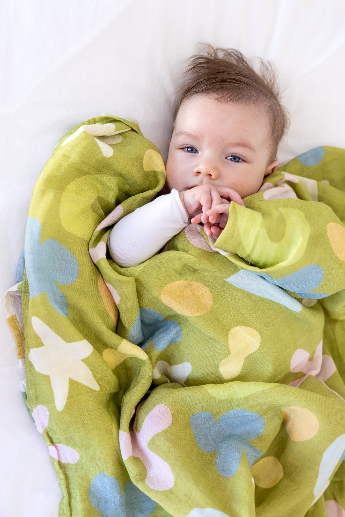 A young child with blue eyes and light brown hair lying in bed, wrapped in a green blanket with a colorful butterfly and polka dot pattern.