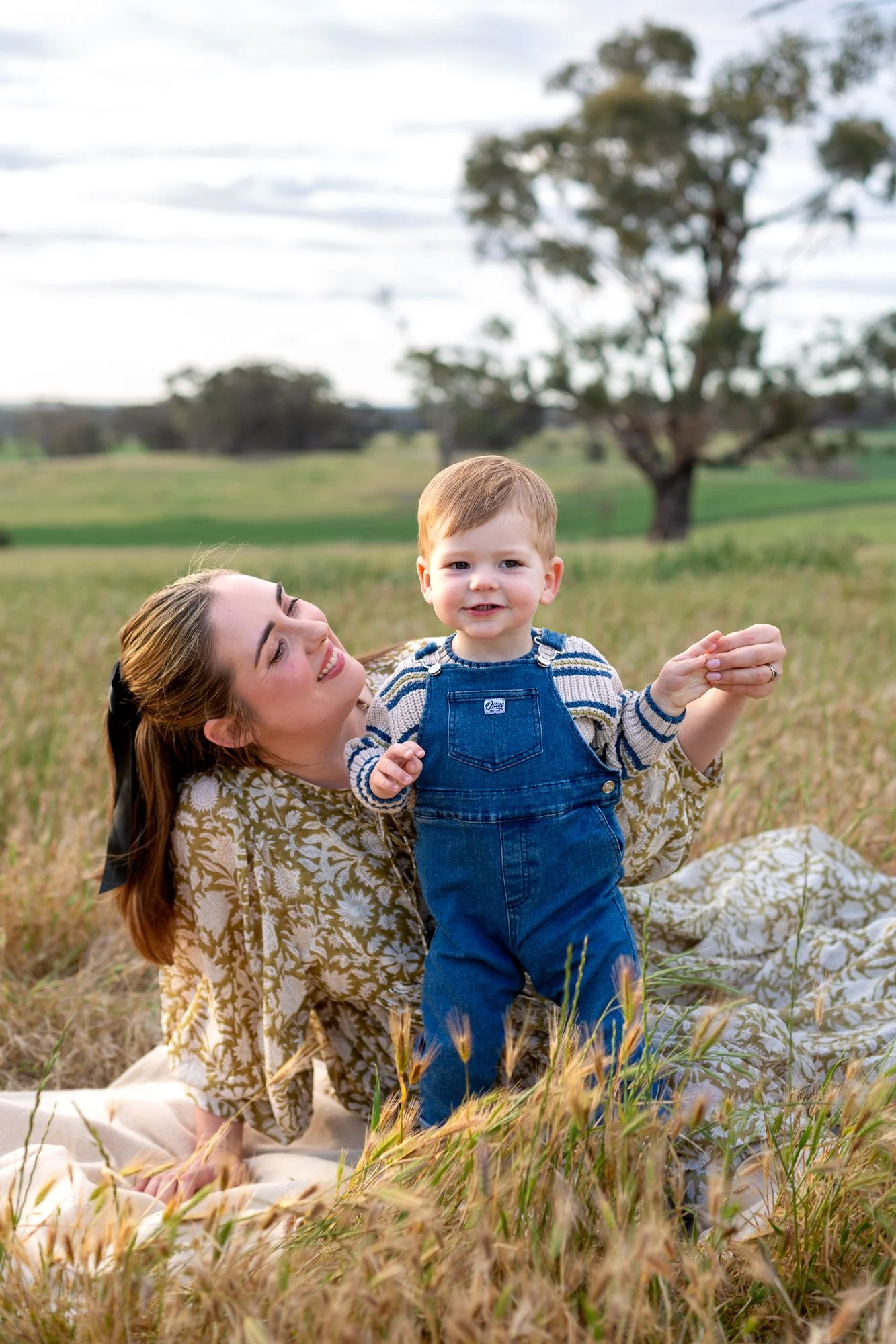 A woman with a black ribbon in her hair lying on a blanket in a grassy field, holding a young boy in blue overalls and a striped shirt, with a tree and open landscape in the background.