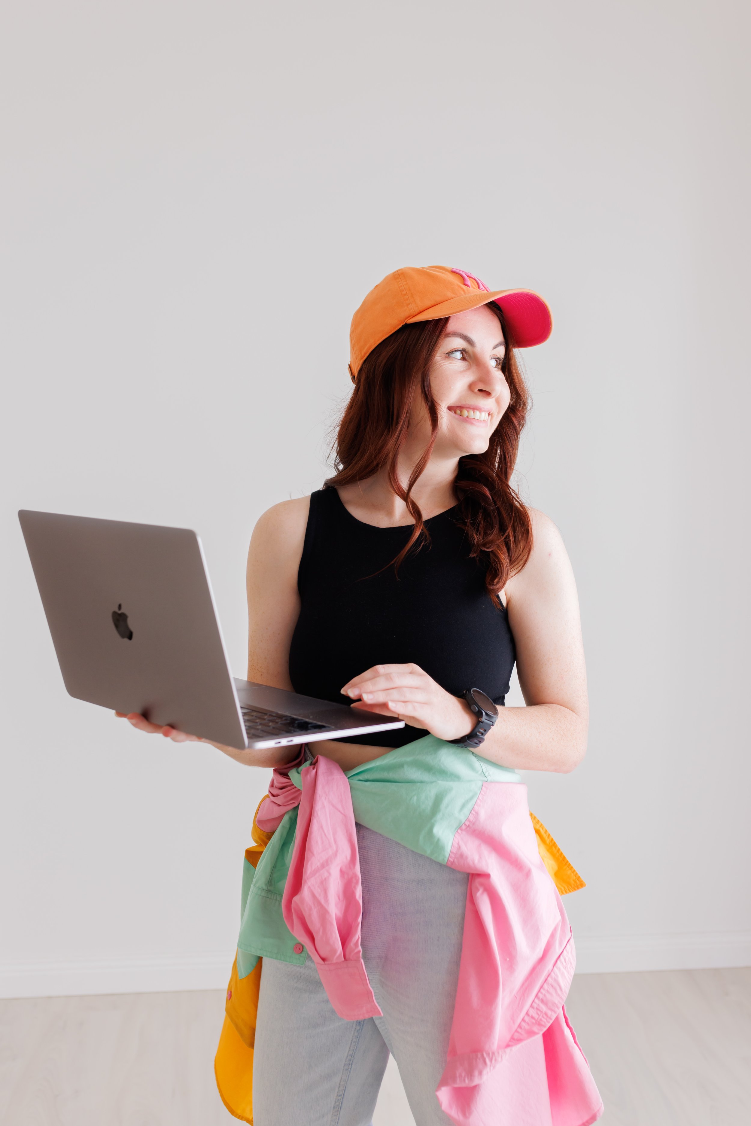 A young woman with red hair and light skin holding a silver MacBook in her right hand, smiling and looking to her right. She is wearing a black sleeveless top, a multicolored jacket tied around her waist, an orange and pink cap, a black smartwatch, and standing against a plain light gray background.