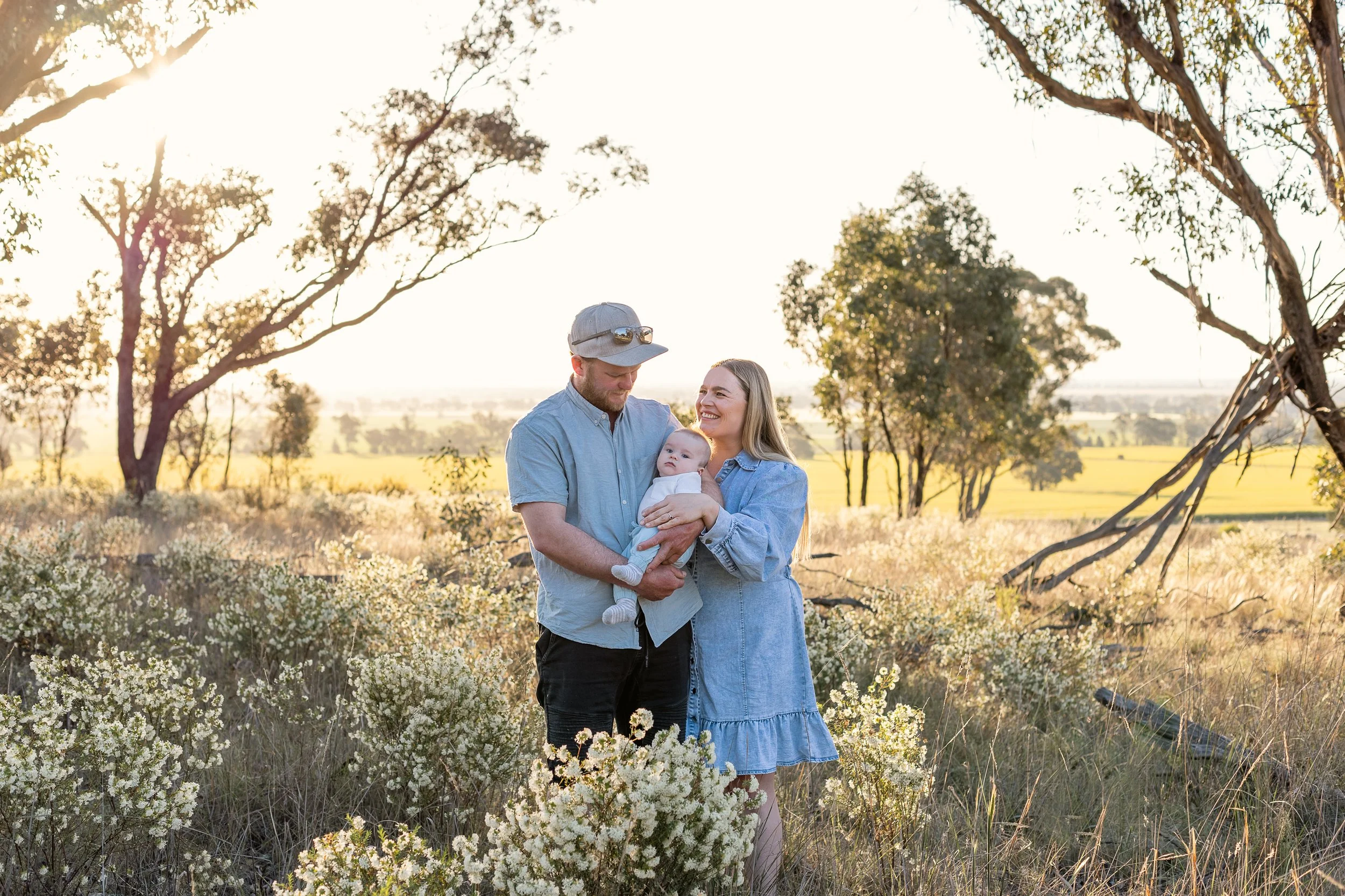 A family of three, a man, woman, and baby, standing in a sunlit field with trees and open land in the background, enjoying a moment together.