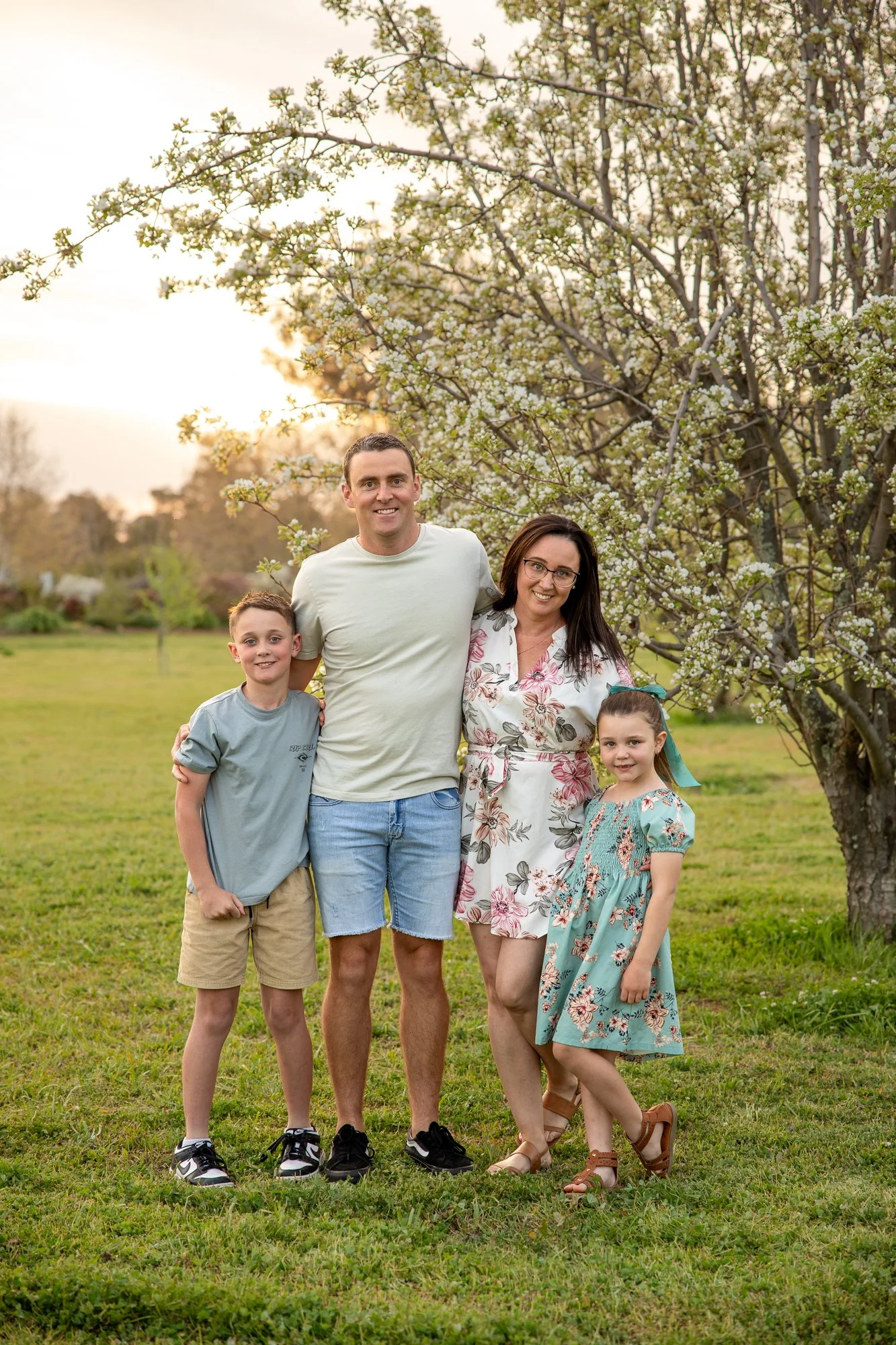 A family of four outdoors near a blooming tree at sunset. The man and woman are standing close together, with the children beside them. The boy is wearing a gray t-shirt and beige shorts, and the girl is wearing a light blue floral dress. The backgro