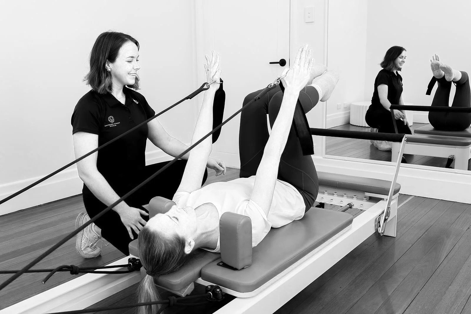 A physical therapy session with a woman lying on a reformer Pilates machine, using resistance bands with assistance from a therapist, in a therapy studio with a mirror and another patient in the background.