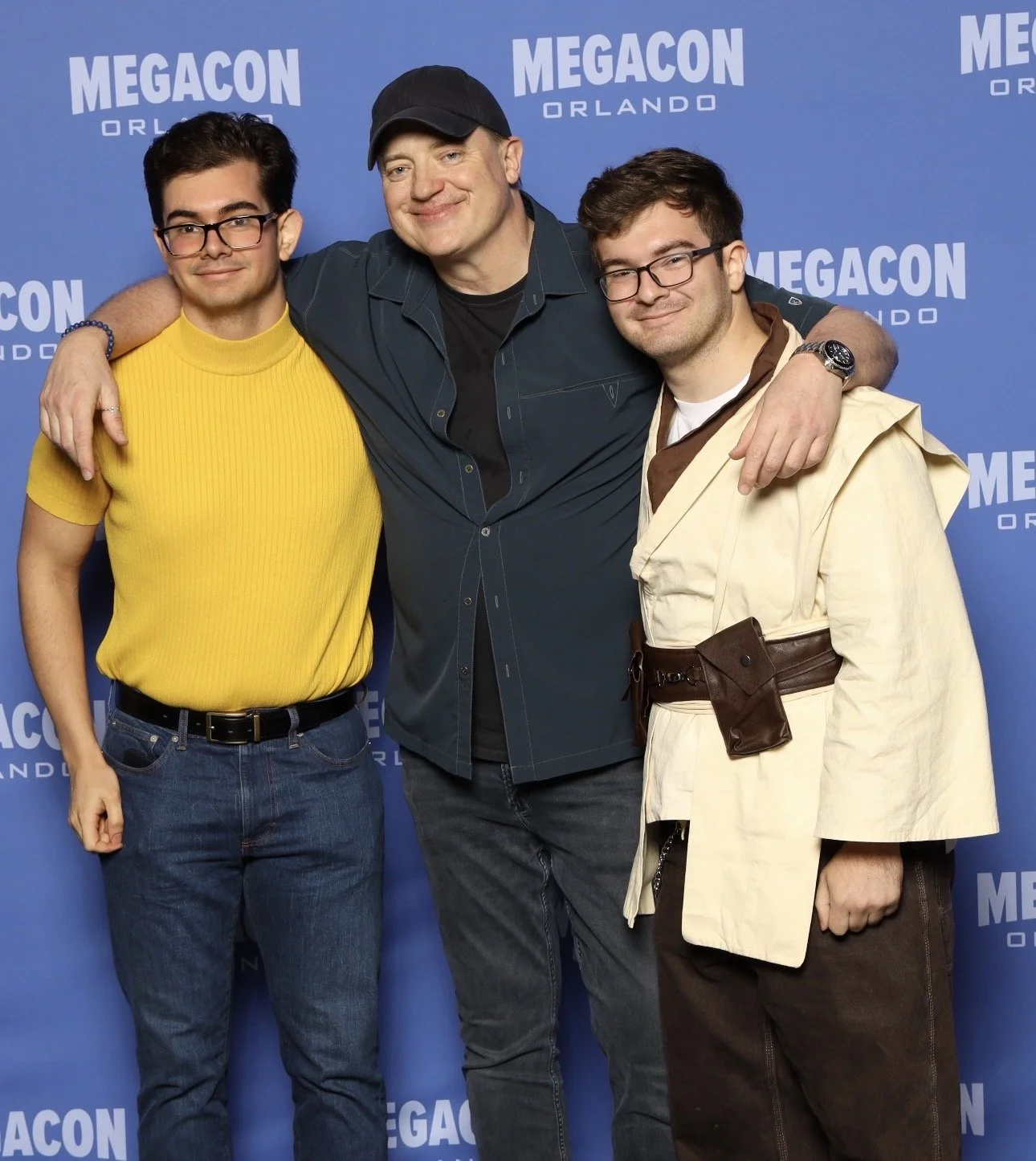 Three men standing together with their arms around each other in front of a blue backdrop with the text 'MEGACON ORLANDO'. They are smiling and looking at the camera.