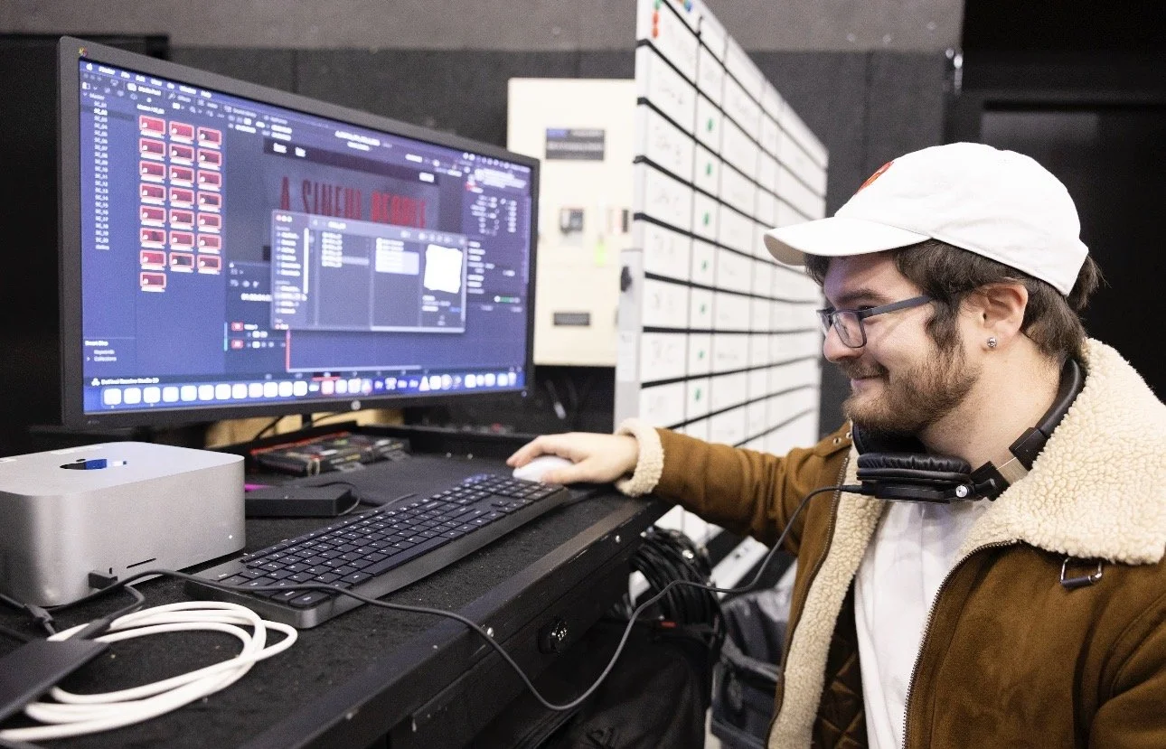 A man with glasses, a white cap, and earphones around his neck is working on a computer with a large monitor. The monitor displays a digital audio workstation (DAW) interface with tracks and editing tools. He is smiling and using a mouse with his rig