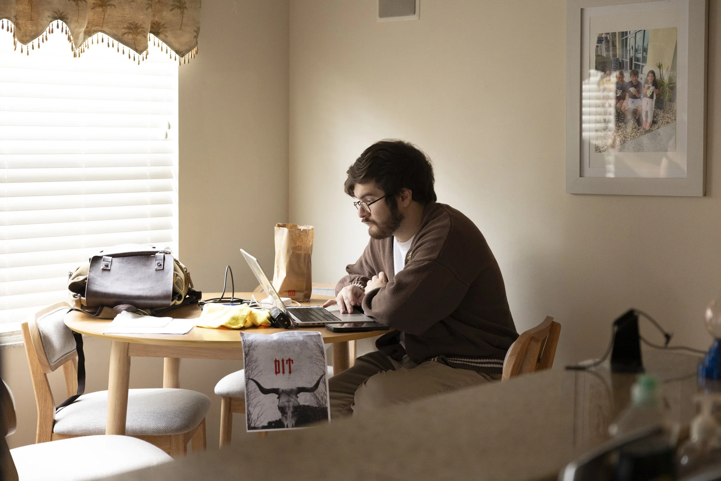A man with glasses and a beard working on a laptop at a round wooden table in a room with beige walls and a window with blinds. There are various items on the table, including a black bag, a beige bag, a brown paper bag, a yellow cloth, and some pape