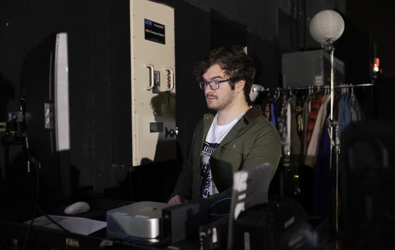 A young man with dark hair and glasses working on a computer in a dark room that appears to be a studio or backstage area, with clothing on a rack and a floor lamp in the background.